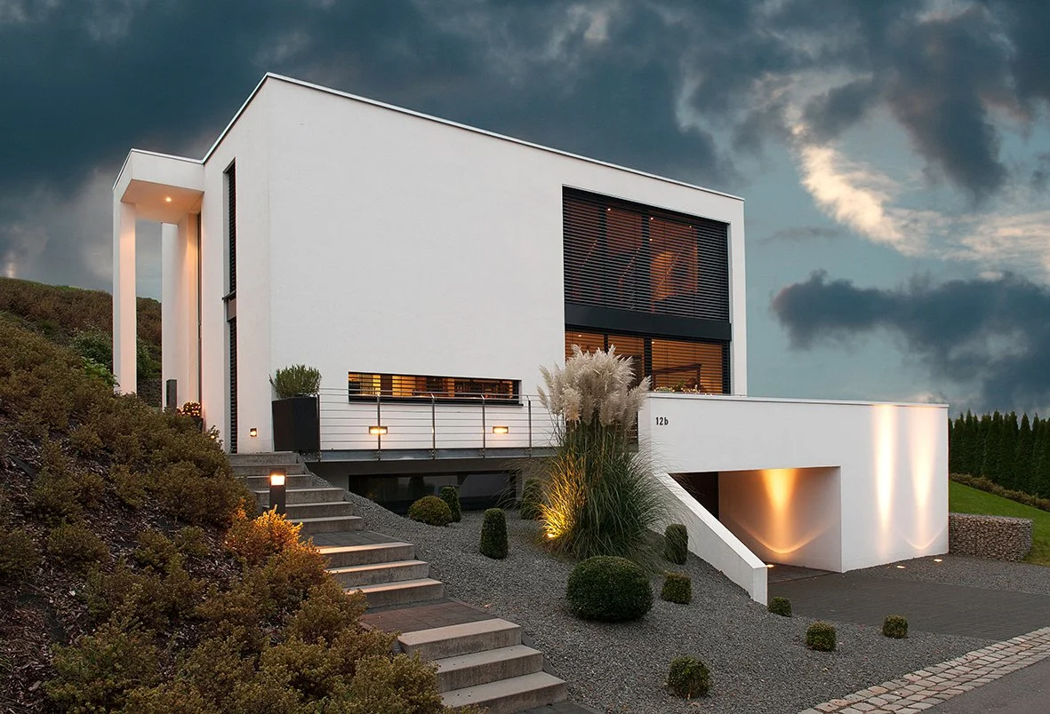 Modernes weißes Haus auf einem Hügel mit Treppen und gepflegtem Garten bei Sonnenuntergang.