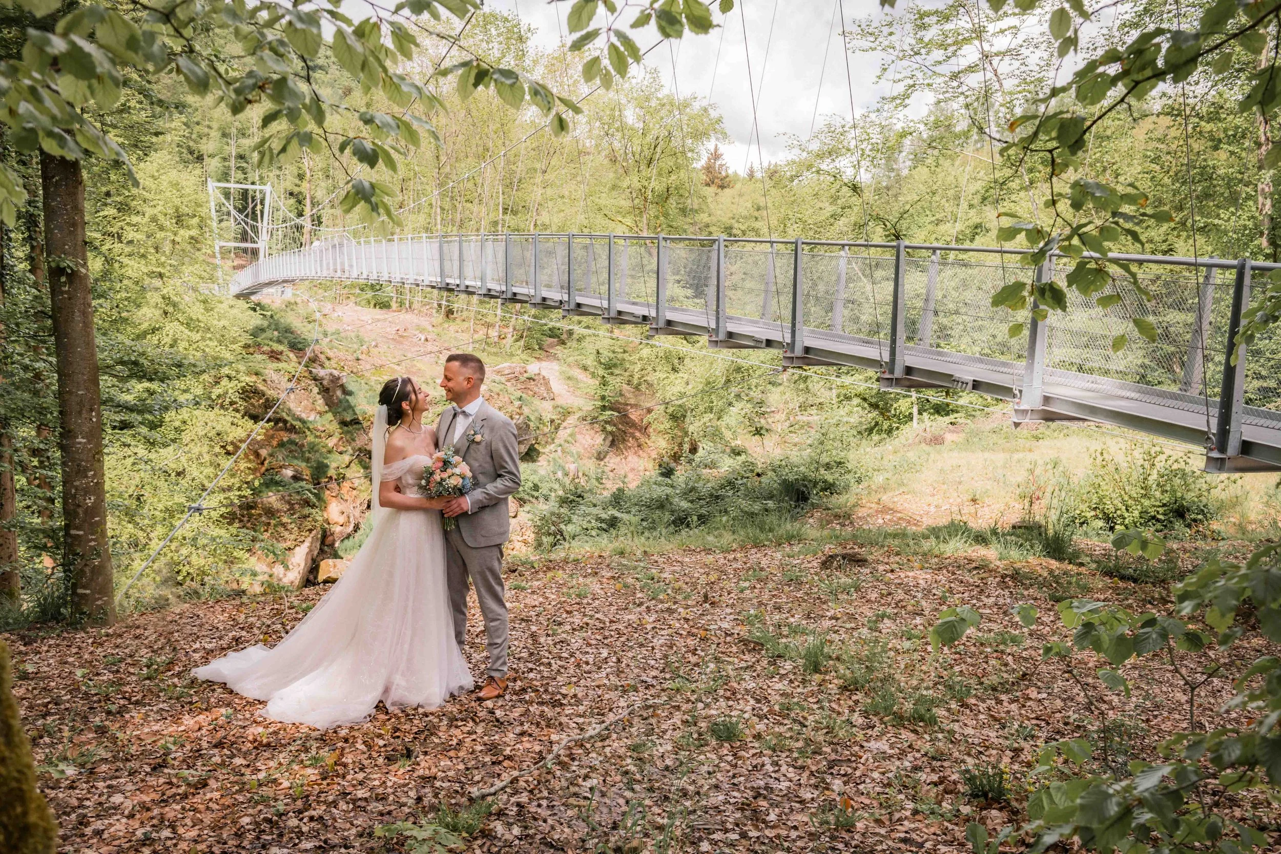 Ein Brautpaar im Wald, die Frau in einem weißen Brautkleid mit Schleier, der Mann in einem grauen Anzug, halten sich gegenüber, umgeben von Bäumen und Blättern, mit einer Hängebrücke im Hintergrund.