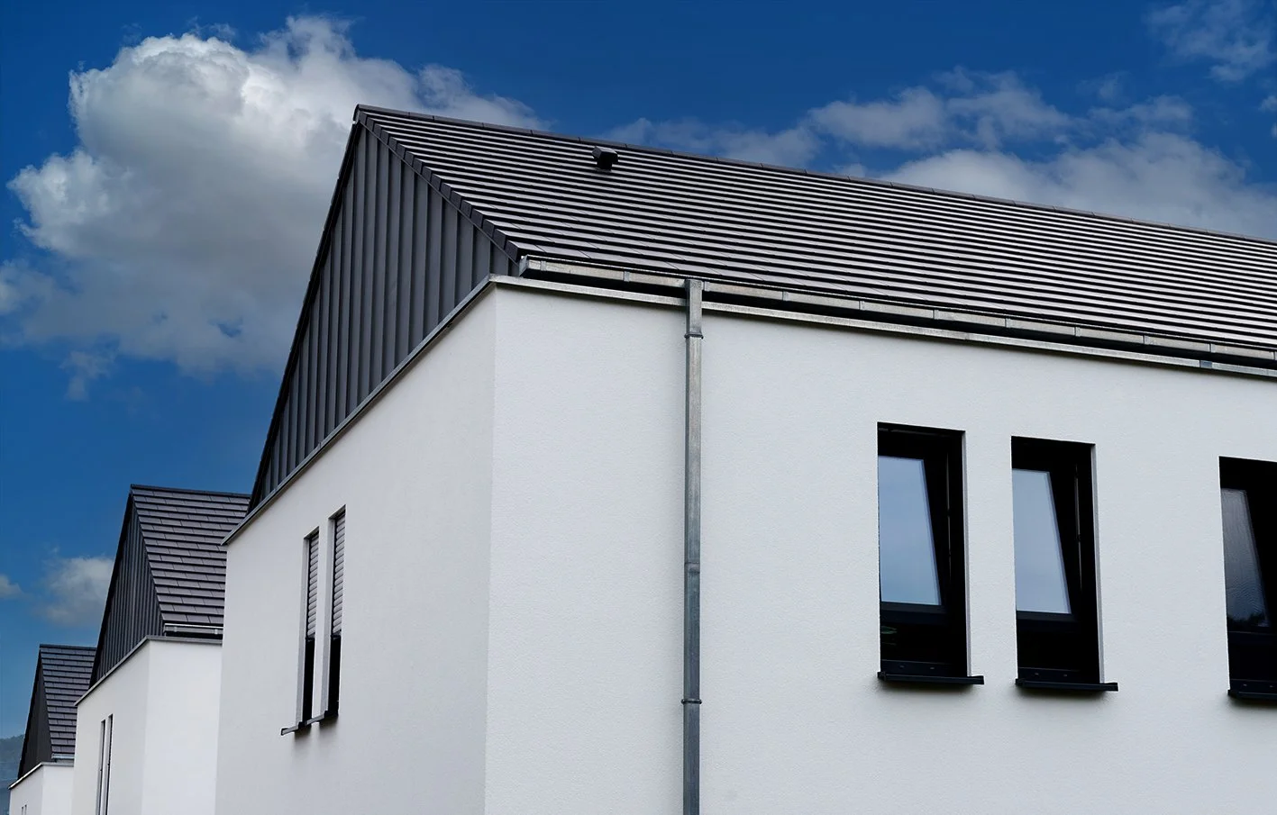 Moderne weiße Mehrfamilienhäuser mit schwarzen Fenstern und dunklem Satteldach gegen blauen Himmel mit Wolken.
