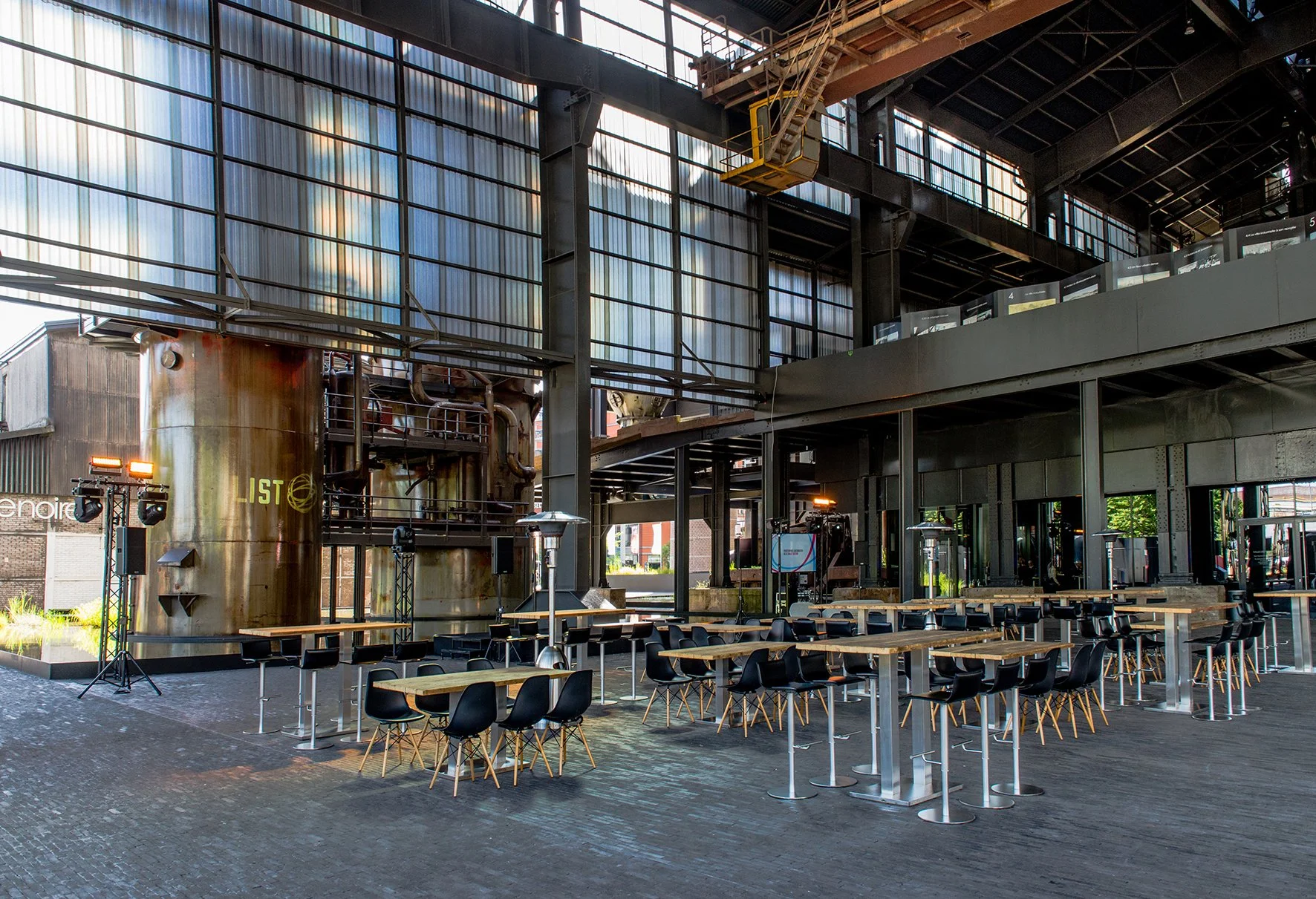 Innenansicht eines modernen Veranstaltungsortes mit mehreren Tischen und schwarzen Stühlen, großen Fenstern, einer schwarzen Metallkonstruktion und einer weißen Wand mit Lichtinstallationen