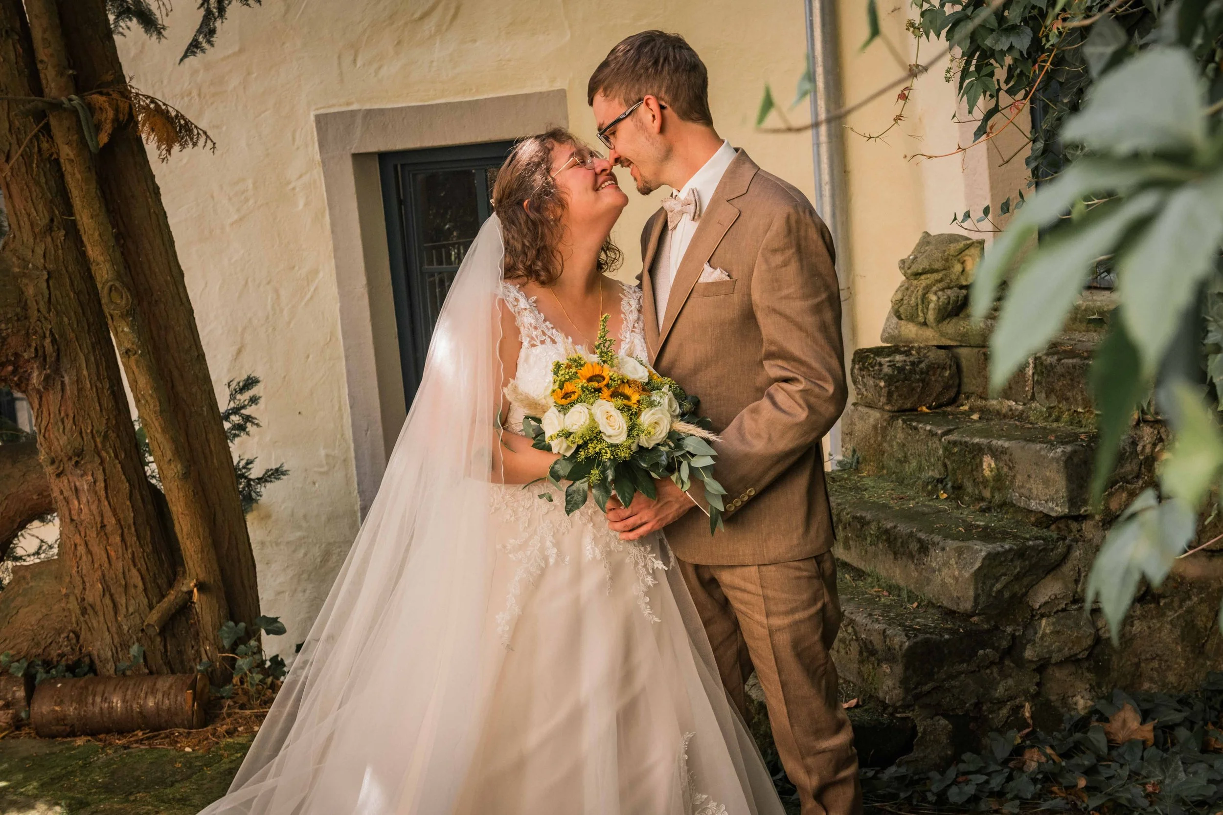 Ein Brautpaar bei ihrer Hochzeit, die Braut trägt ein weißes Hochzeitskleid und einen Schleier, der Bräutigam trägt einen braunen Anzug, sie stehen sich gegenüber und lächeln sich an, die Braut hält einen Blumenstrauß, im Hintergrund eine alte Hauswa