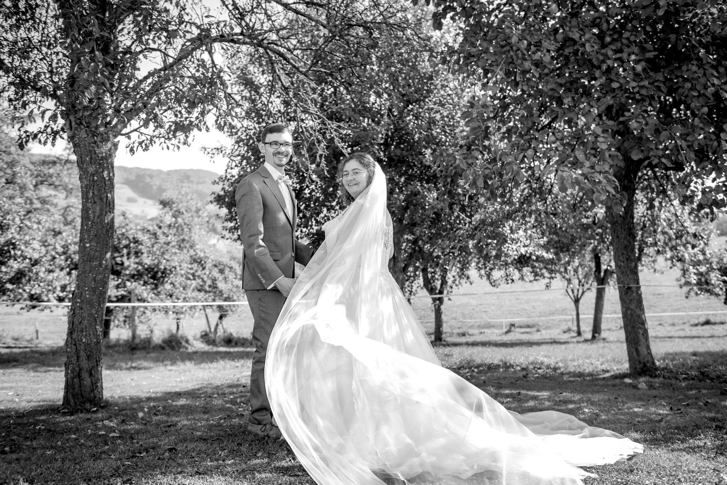 Ein Brautpaar steht im Freien unter Bäumen, die Braut trägt ein langes Brautkleid mit Schleier, der Bräutigam trägt einen Anzug, beide lächeln.