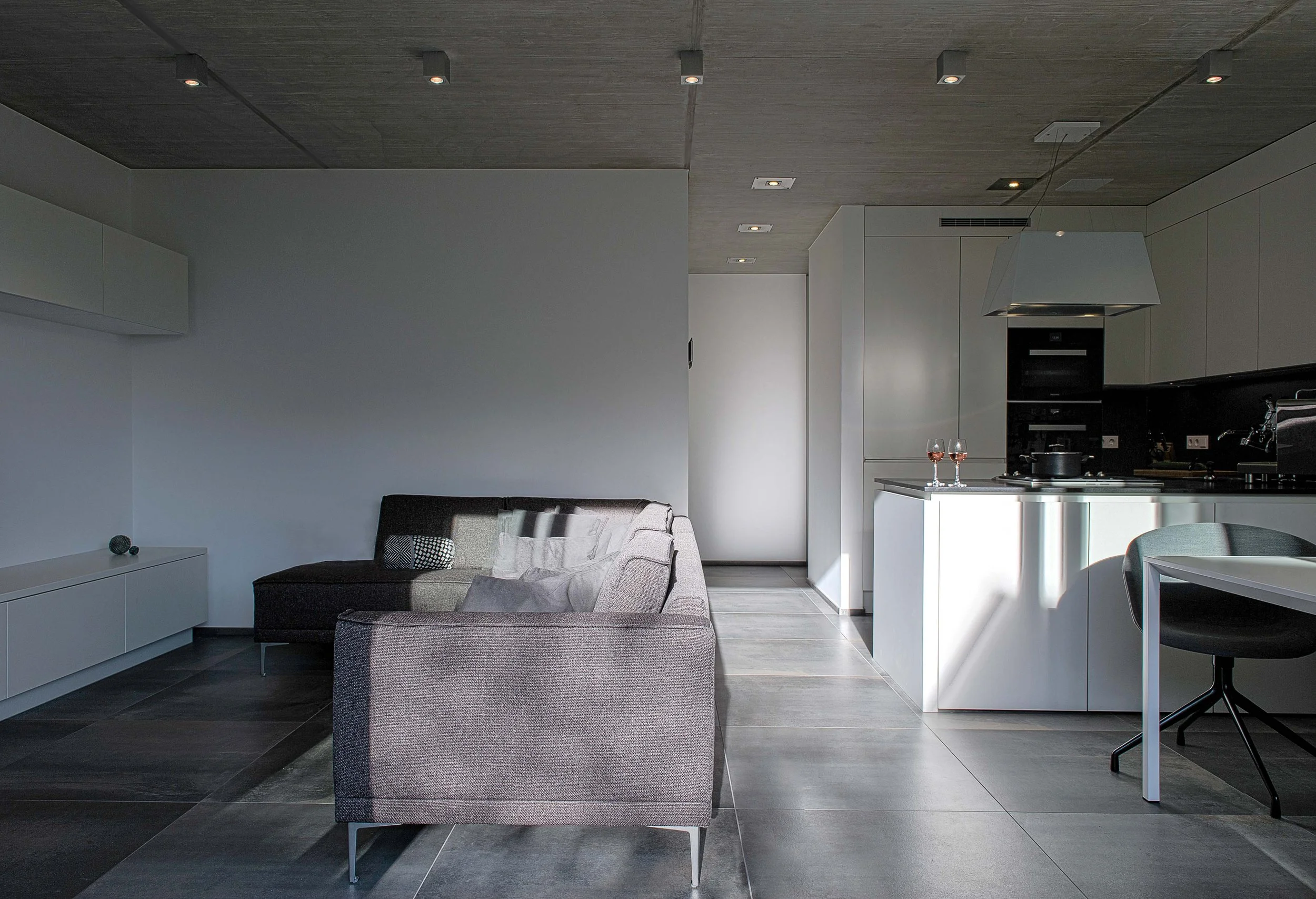 Moderner offen gestalteter Wohn- und Küchenbereich, mit grauer Couch, dunklem Boden und weißem Küchenblock, zwei Weingläser auf der Kochinsel, schwarze Einbaugeräte.
