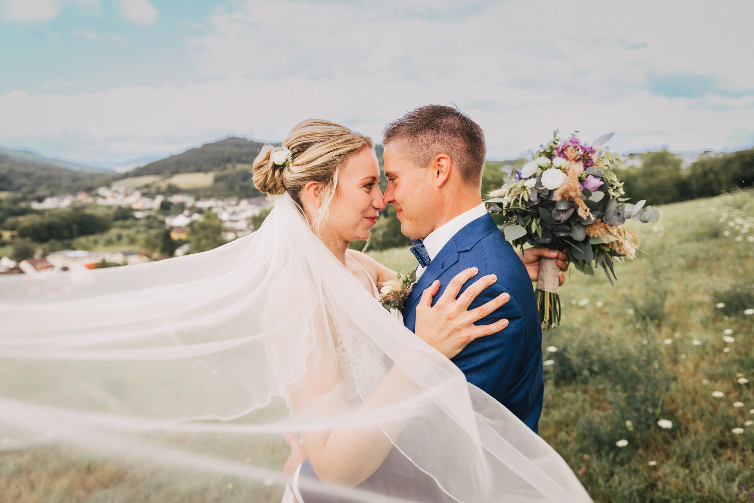 Ein Brautpaar im Hochzeitsoutfit, das sich liebevoll berührt, im Freien vor einer hügeligen Landschaft, die mit Häusern und Bäumen bedeckt ist. Die Braut trägt ein weißes Kleid und einen Schleier, der im Wind weht, der Bräutigam trägt einen blauen An