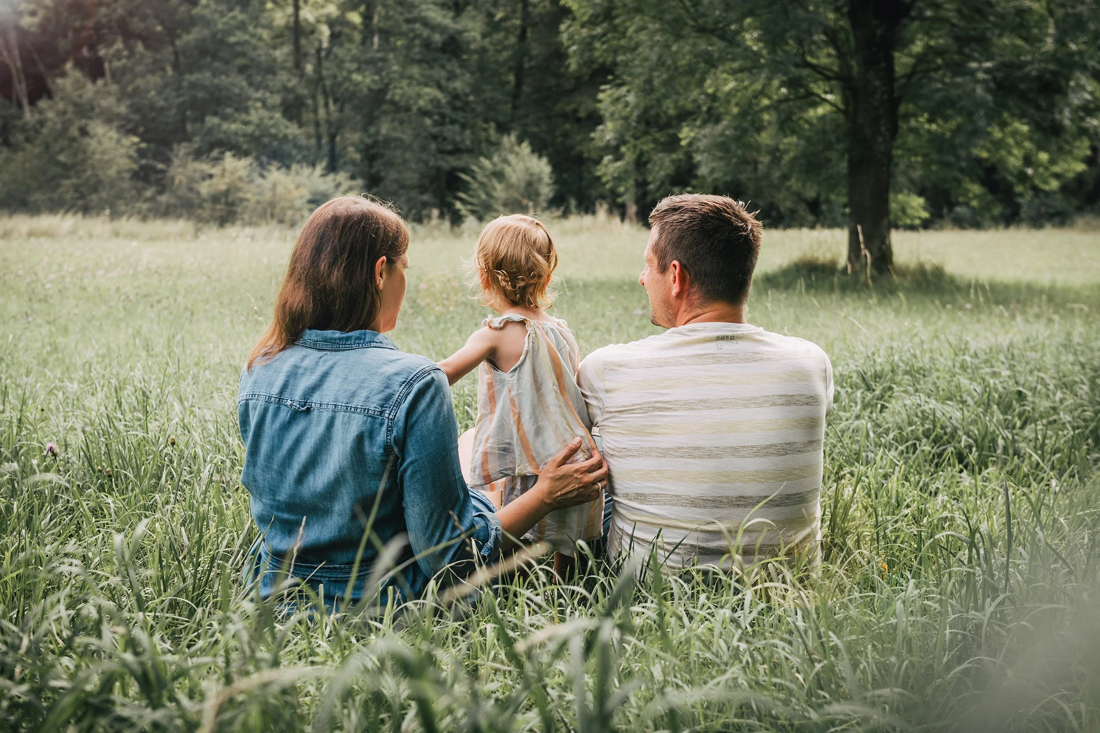 Familie mit einem Kind sitzt auf einer Wiese im Park, umgeben von Bäumen, und genießt die Natur.