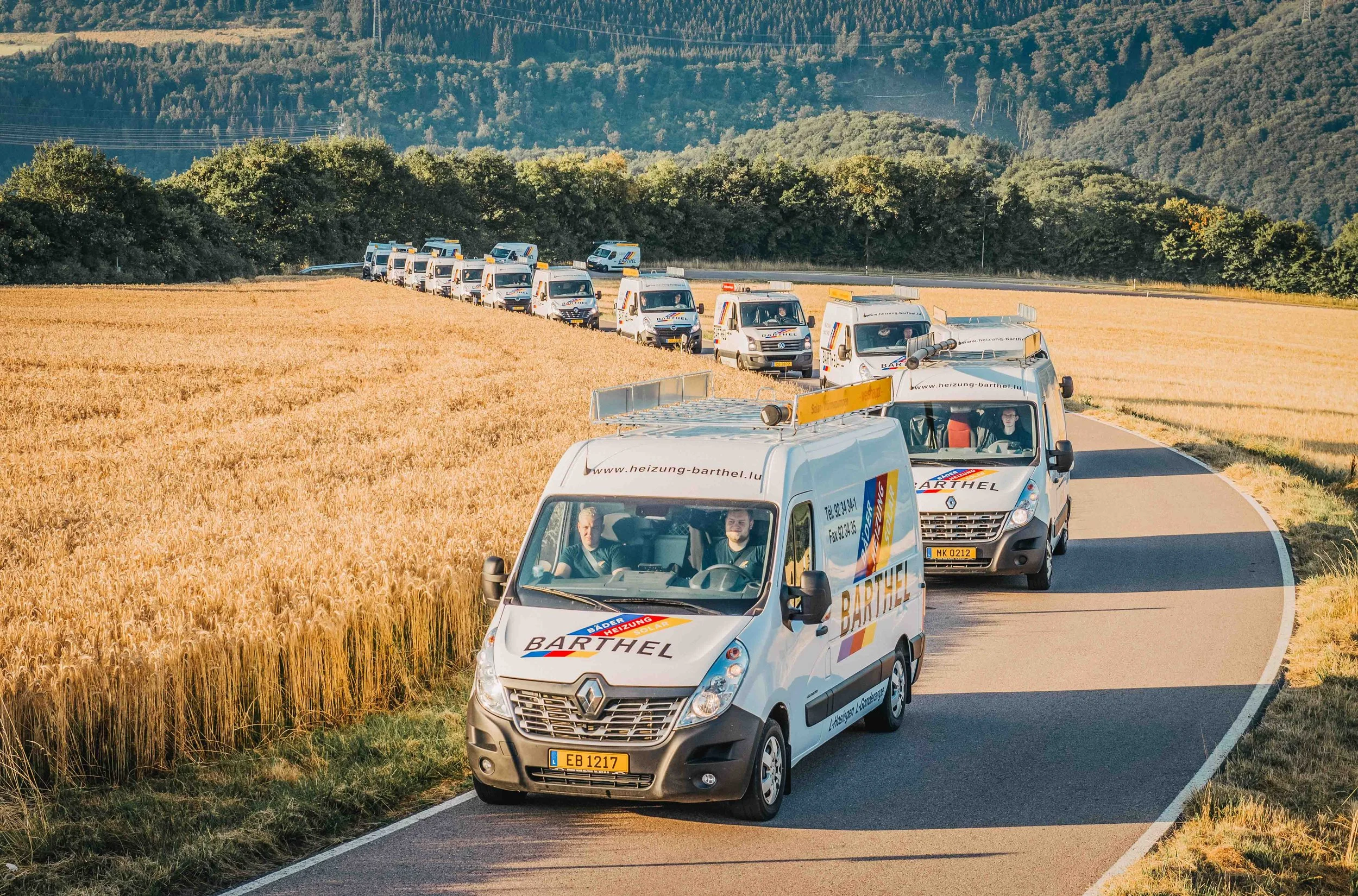 Mehrere weiße Firmenfahrzeuge mit Werbeaufdruck fahren auf einer Landstraße neben einer goldgelben Getreidefeld, umgeben von grünen Hügeln und Wäldern, bei Sonnenuntergang.