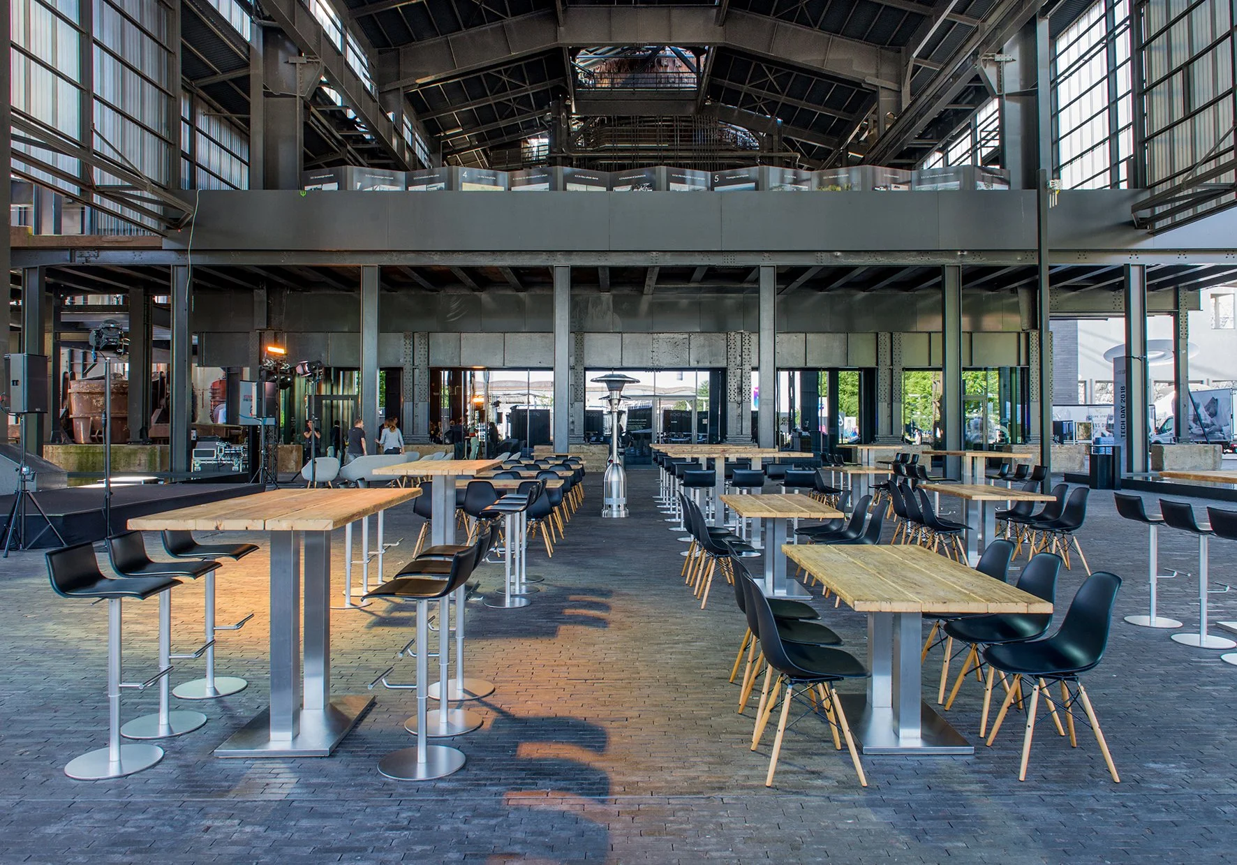 Innenansicht eines modernen, offenen Veranstaltungsraums mit hohen Decken, Holztischen und schwarzen Stühlen, viel natürlichem Licht durch große Fenster, und jemandem, der an der Seite arbeitet.
