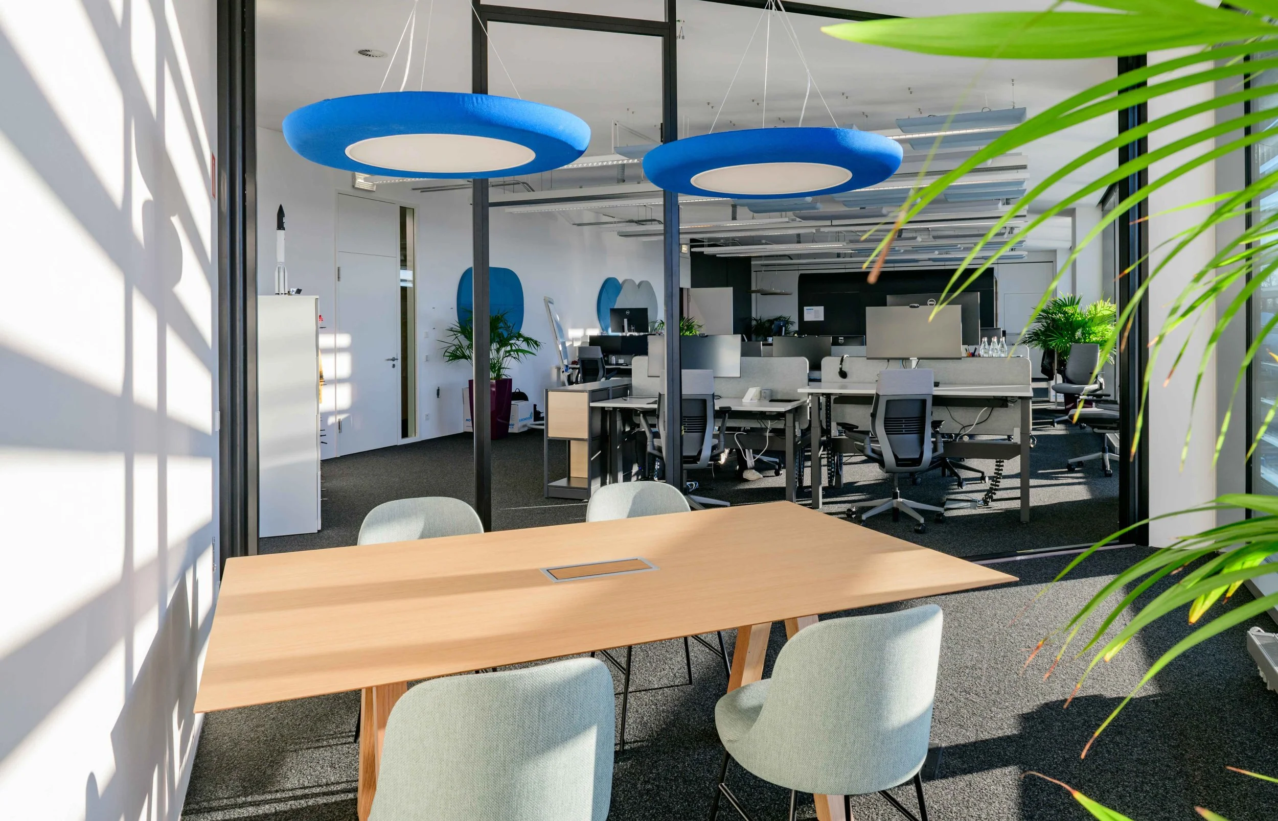 Ein moderner, heller Büroraum mit einem Holztisch und sechs Stühlen im Vordergrund. Im Hintergrund befinden sich Arbeitsplätze mit Stühlen, Pflanzen und technischen Geräten. Zwei große blaue Deckenleuchten hängen von der Decke.
