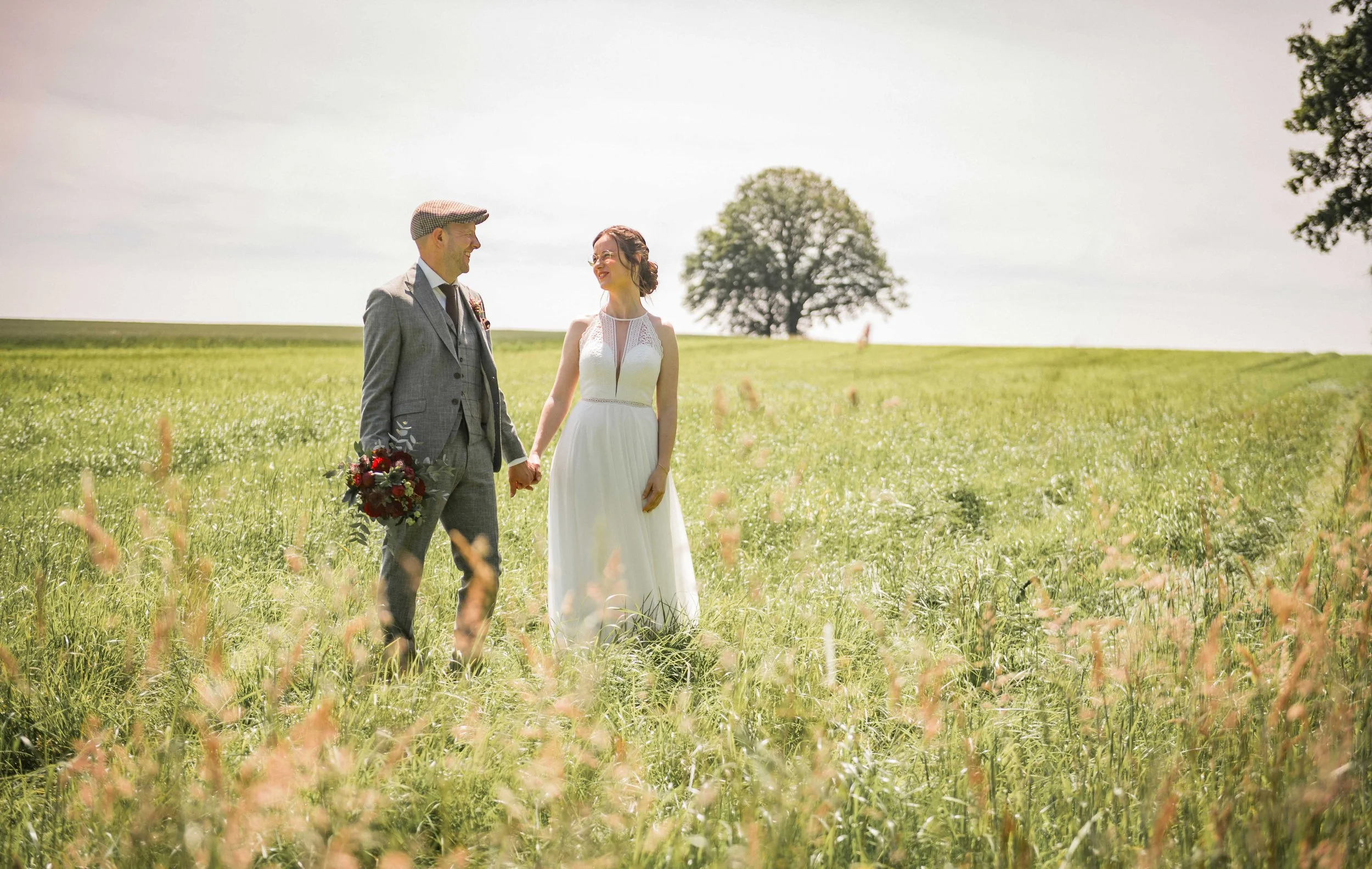 Ein Paar in Hochzeitskleidung in einer grünen Wiese, Hand in Hand, bei Sonnenschein, mit einem einzelnen Baum im Hintergrund.