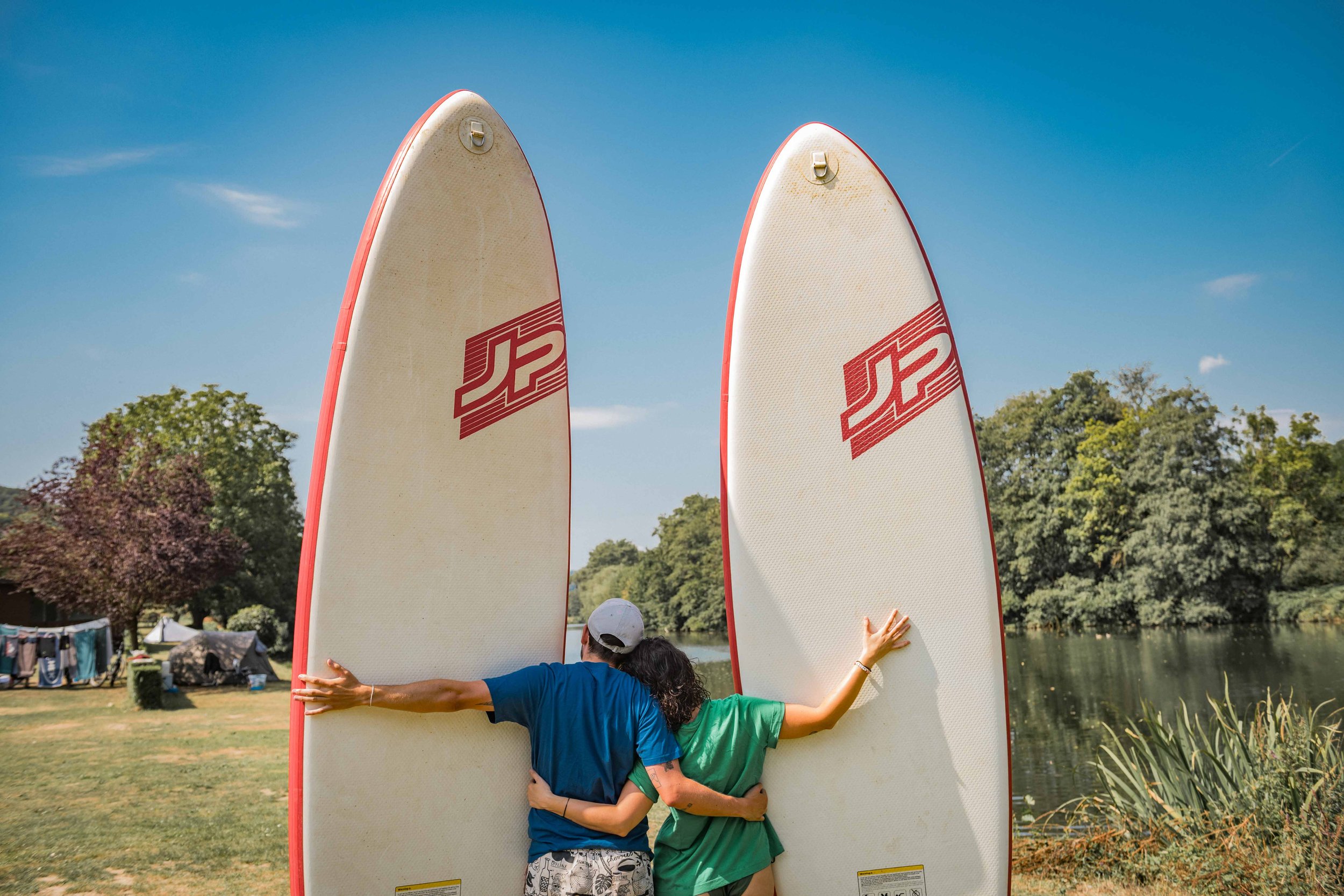 Zwei Personen umarmen sich vor einem See, die eine Person hält zwei große Stand-up-Paddle-Boards skyward.