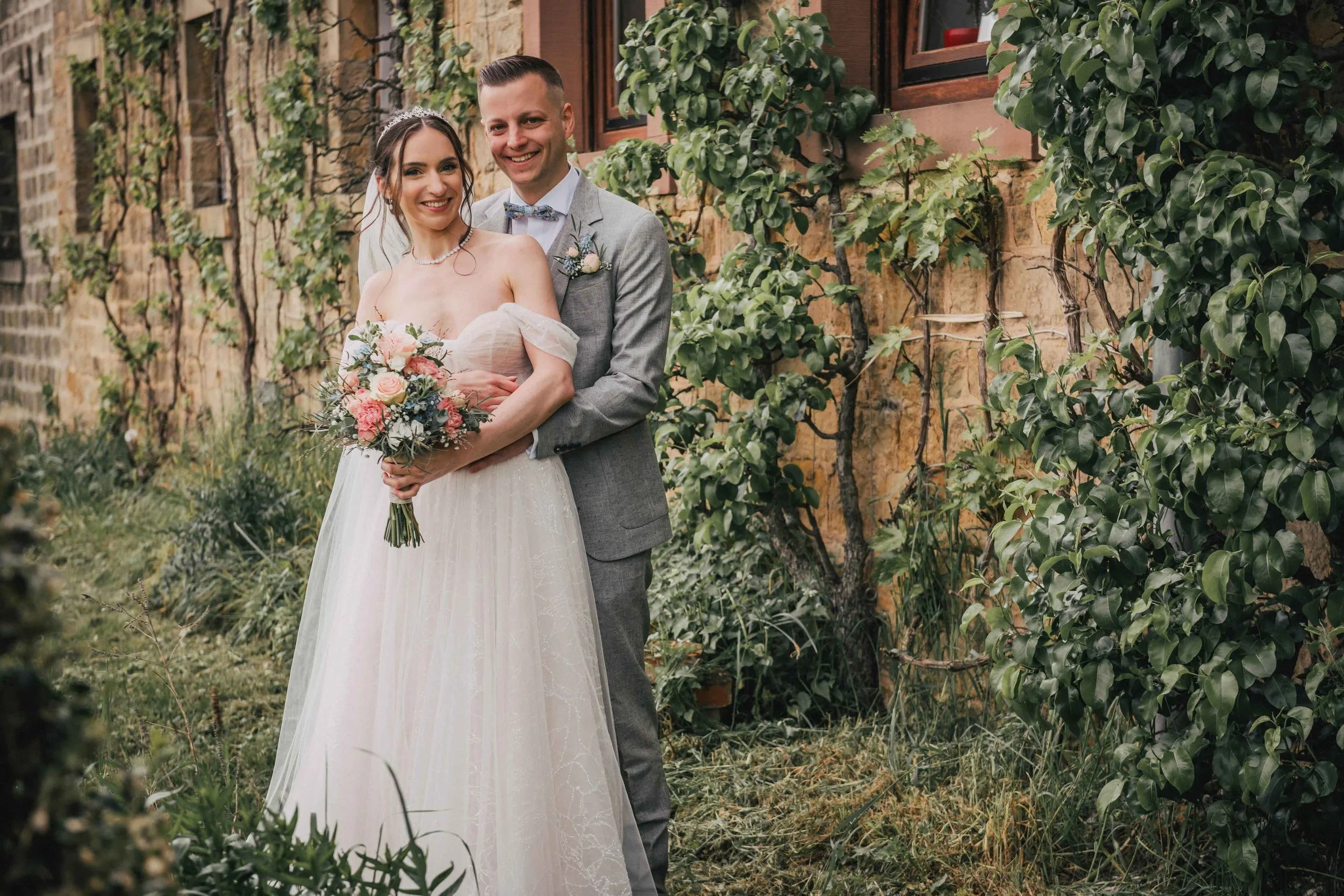 Ein Brautpaar steht draußen vor einer Steinmauer, umgeben von grünen Pflanzen. Die Braut trägt ein weißes Hochzeitskleid und hält einen Blumenstrauß. Der Bräutigam trägt einen grauen Anzug und eine Fliege. Sie lächeln in die Kamera.