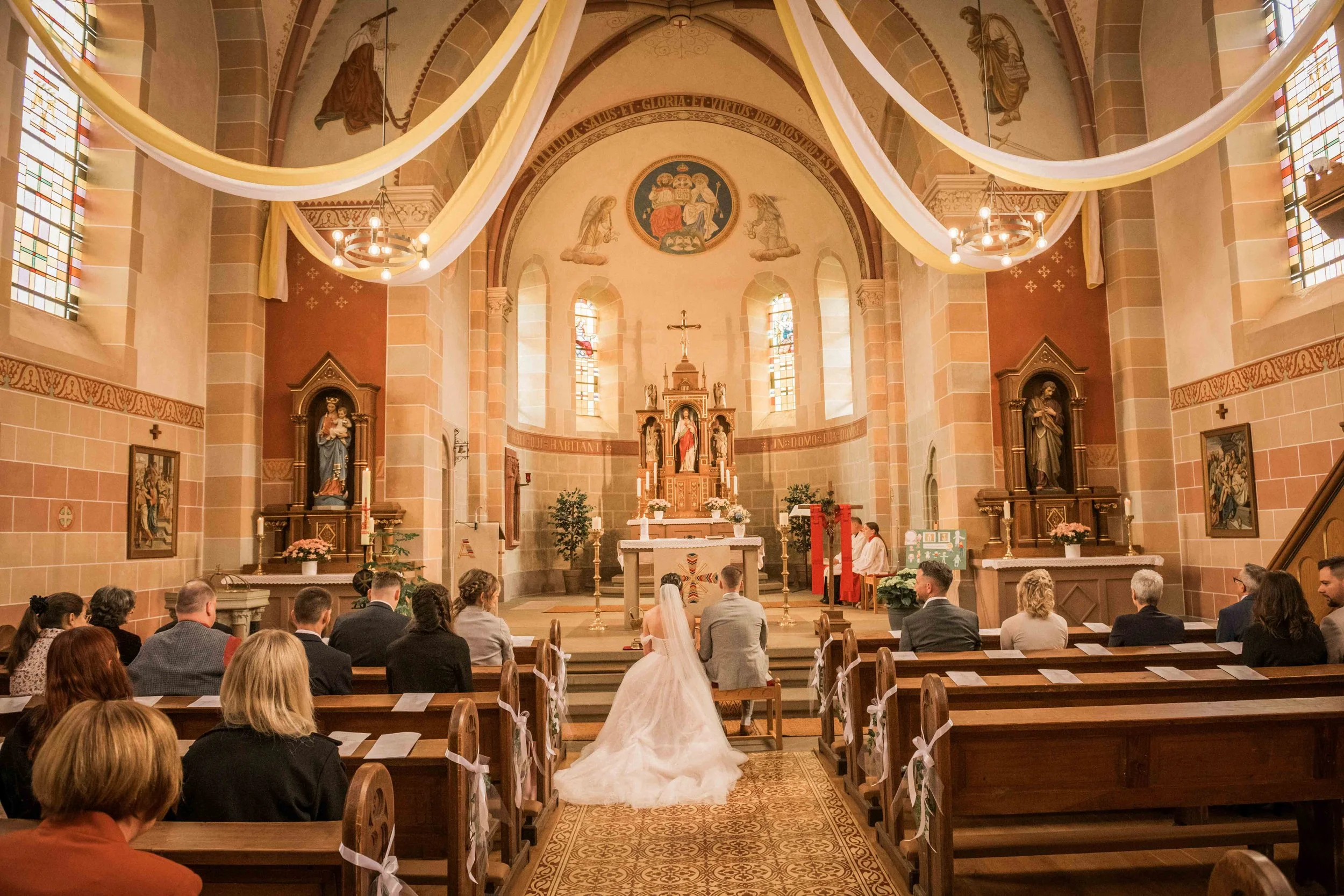 Hochzeitszeremonie in einer Kirche mit Braut und Bräutigam, umgeben von Gästen, im Hintergrund Altar, religiöse Symbole und Kirchenfenster, warme Beleuchtung.