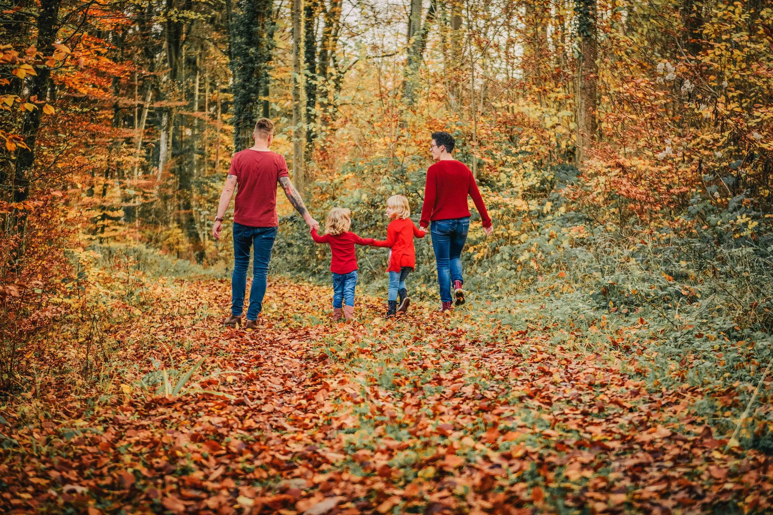 Eine Familie mit zwei Erwachsenen und zwei Kindern, alle in roten Kleidung, spaziert durch einen herbstlichen Wald mit buntem Laub auf dem Boden und in den Bäumen.