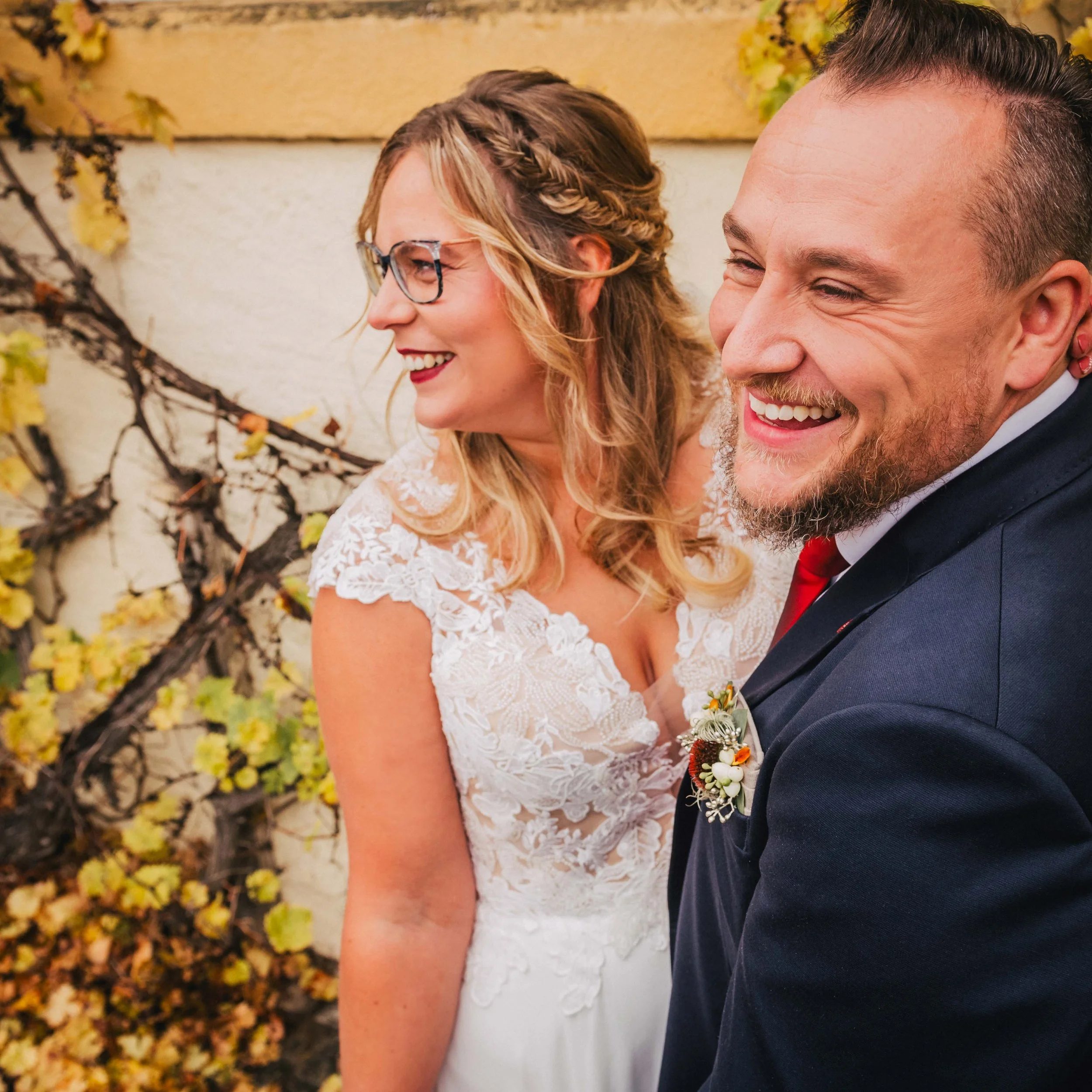 Ein lächelndes Hochzeitspaar, eine Frau in einem weißen Brautkleid mit Brille und Locken, ein Mann in einem dunkelblauen Anzug mit roter Krawatte, vor einer Wand mit Efeu und gelben Blättern.
