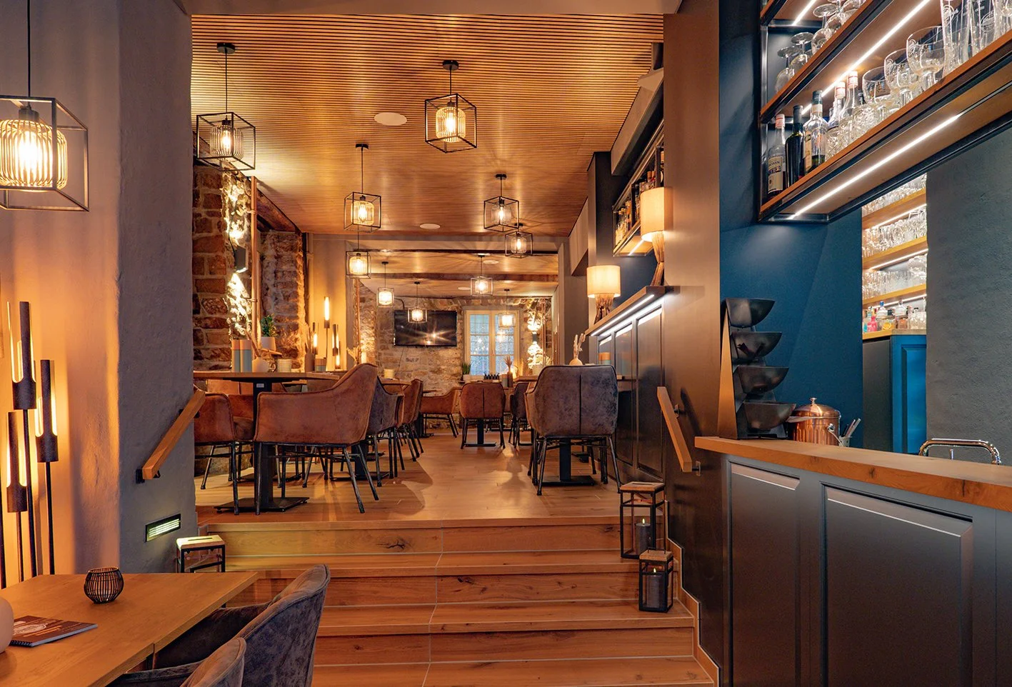 Gemütliches Restaurant mit warmem Licht, Holzdecken und Steinwänden, Tischen und Sessel, dekorative Lampen und ein Fernseher an der Wand.