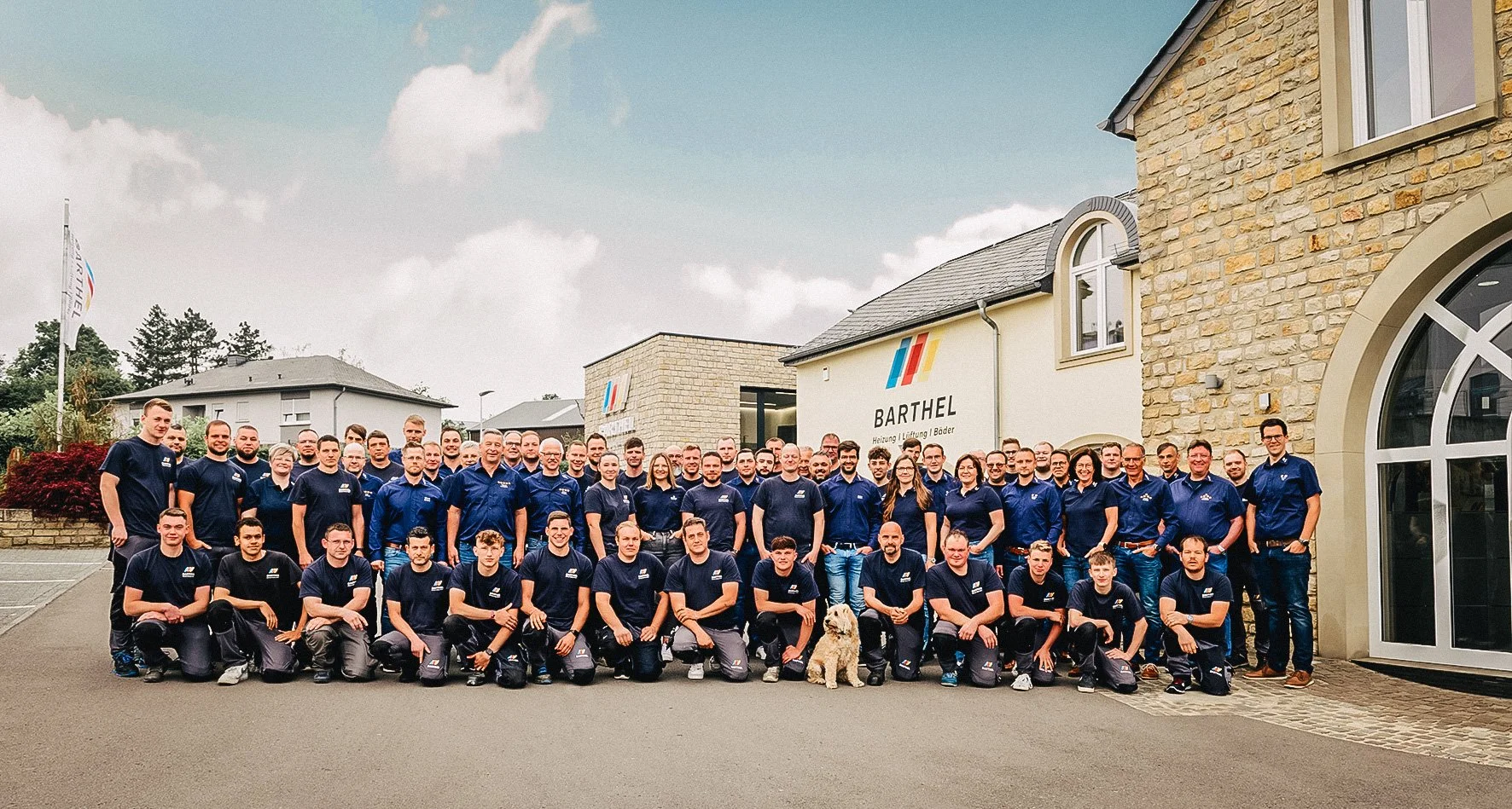 Eine große Gruppe von Mitarbeitenden trägt dunkle Arbeitskleidung mit Firmenlogo und steht vor einem Gebäude mit Firmenbeschriftung "BARTEL" für Heizung, Lüftung und Bäder.