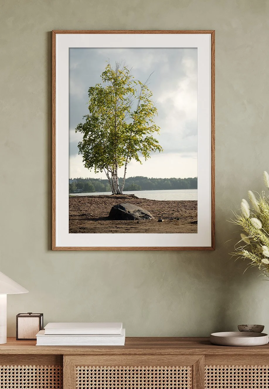 Ein gerahmtes Wandbild zeigt eine einzelne Baum auf einer sandigen Uferfläche mit Wasser im Hintergrund, unter bewölktem Himmel.