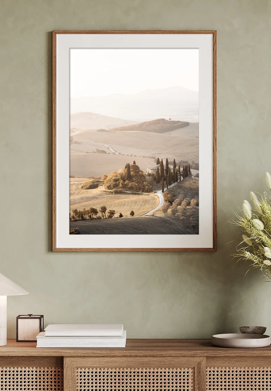 Ein gerahmtes Wandbild zeigt eine ruhige Landschaft mit Hügeln, Zypressen und einer kurvigen Straße in Tönen von Beige und Braun, aufgehängt an einer grünen Wand. Motiv befindet sich in der Toskana, Italien