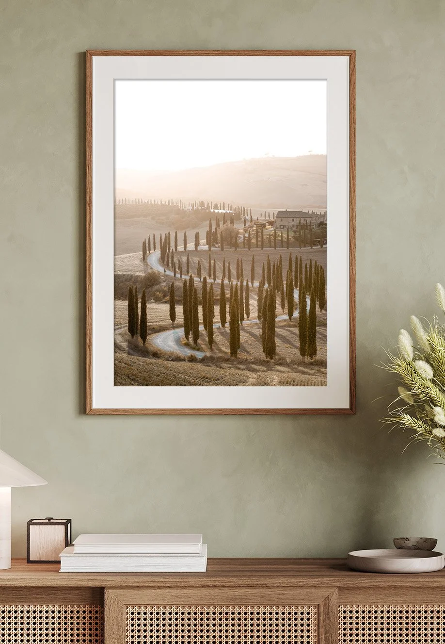 Ein Rahmen mit einem Wandbild eines ländlichen italienischen Hügellands mit einer kurvigen Straße und Zypressenbäumen bei Sonnenuntergang, das an einer Wand in einem Wohnzimmer hängt. Motiv befindet sich in der Toskana, Italien