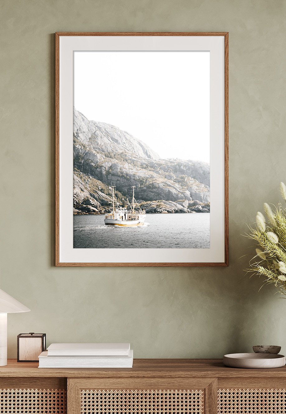 Fotografie eines Bootes auf dem Wasser vor einer felsigen Berglandschaft, an einer weißen Wand in einem Raum aufgehängt. Foto entstand im Nusfjord auf den Lofoten