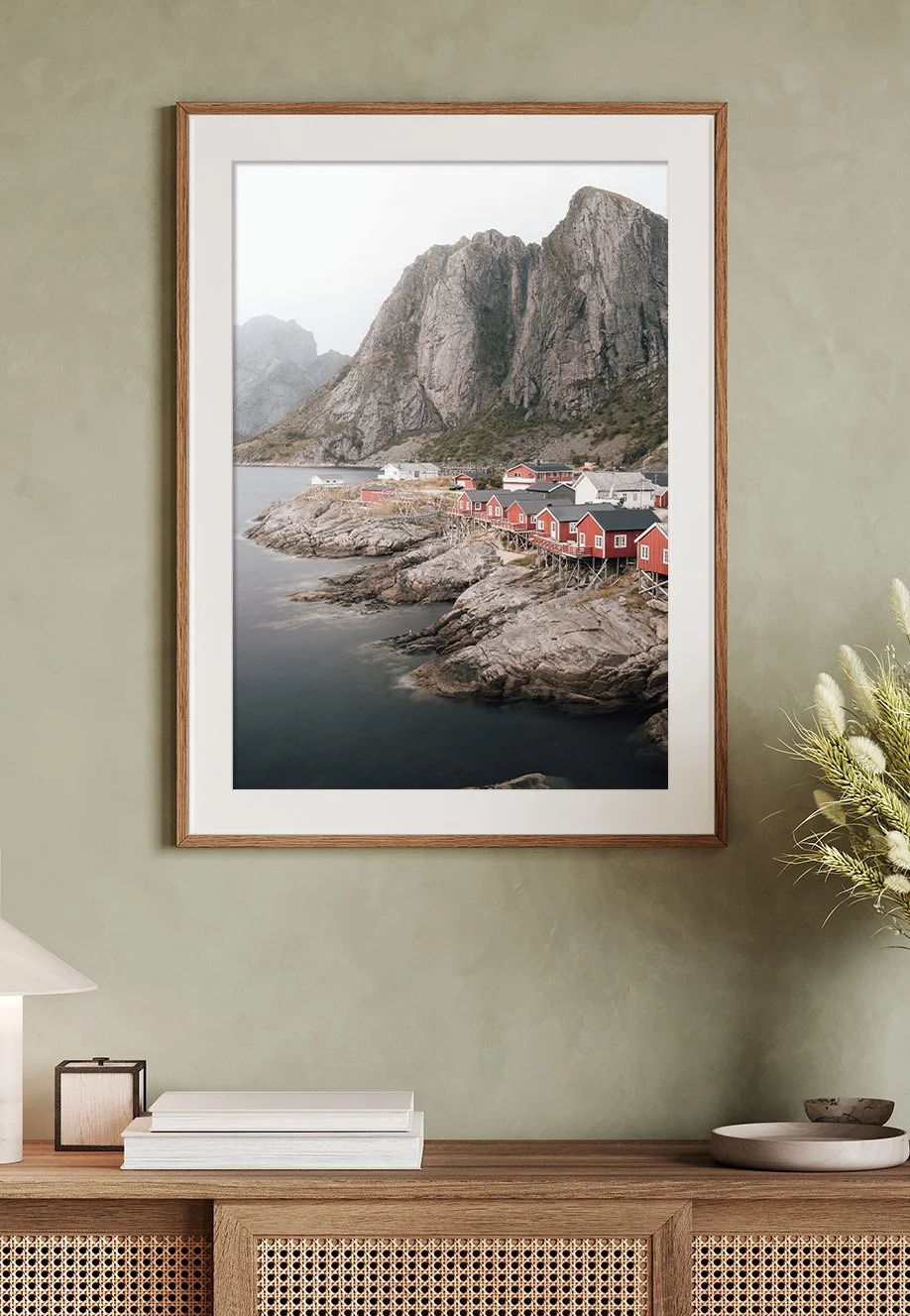 Ein gerahmtes Landschaftsfoto von einer Küstenregion mit roten Holzhäusern, hohen Felsen und Wasser im Vordergrund, an einer grünen Wand in einem Wohnzimmer