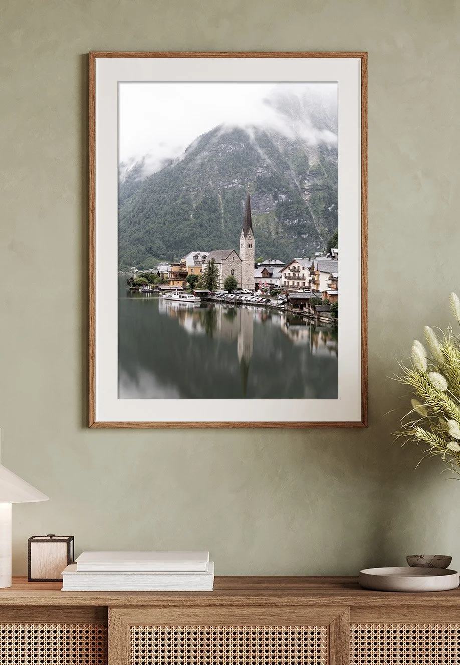 Ein gerahmtes Wandbild zeigt ein Dorf, Hallstatt - Österreich, am Ufer eines Sees mit Bergen im Hintergrund, neblig und bewölkt.