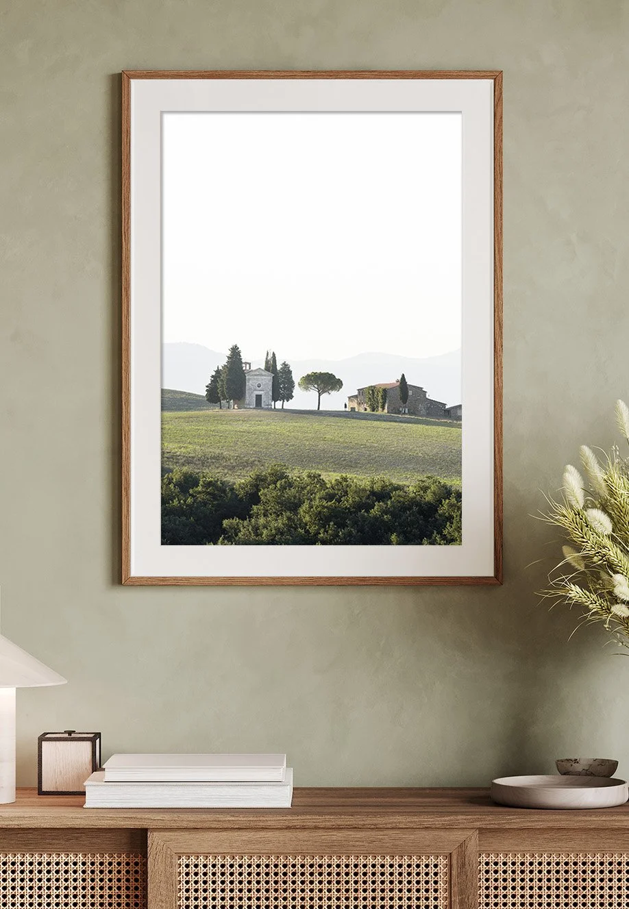 Ein Wandbild eines Landschaftsbildes mit Häusern und Bäumen, eingerahmt und an einer Wand aufgehängt über einem Holzsideboard.