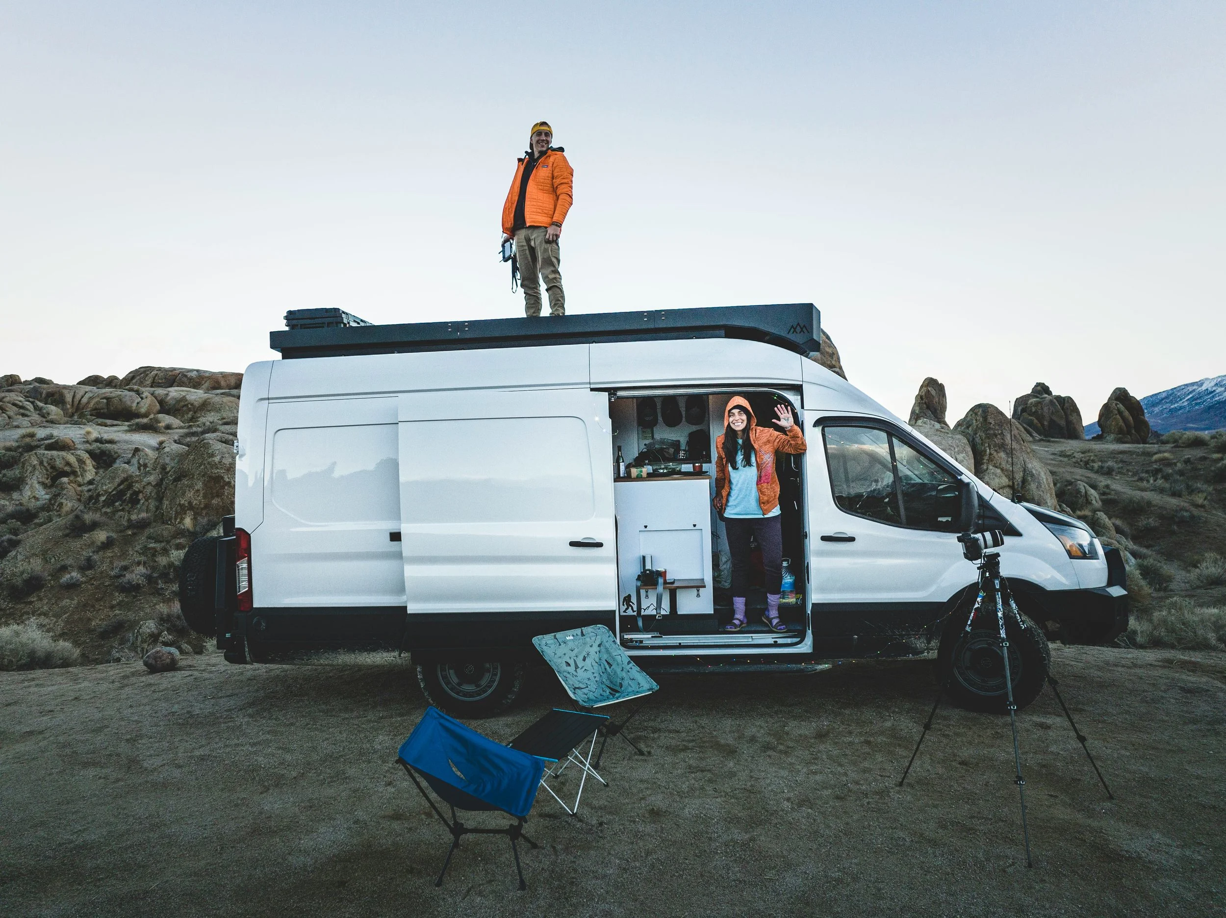 A man and a woman camping outdoors with a white van in a rocky, mountainous area. The woman is inside the van, waving and smiling, while the man is on top of the van, smiling and looking at the camera. Camping chairs and a camera are set up outside the van.