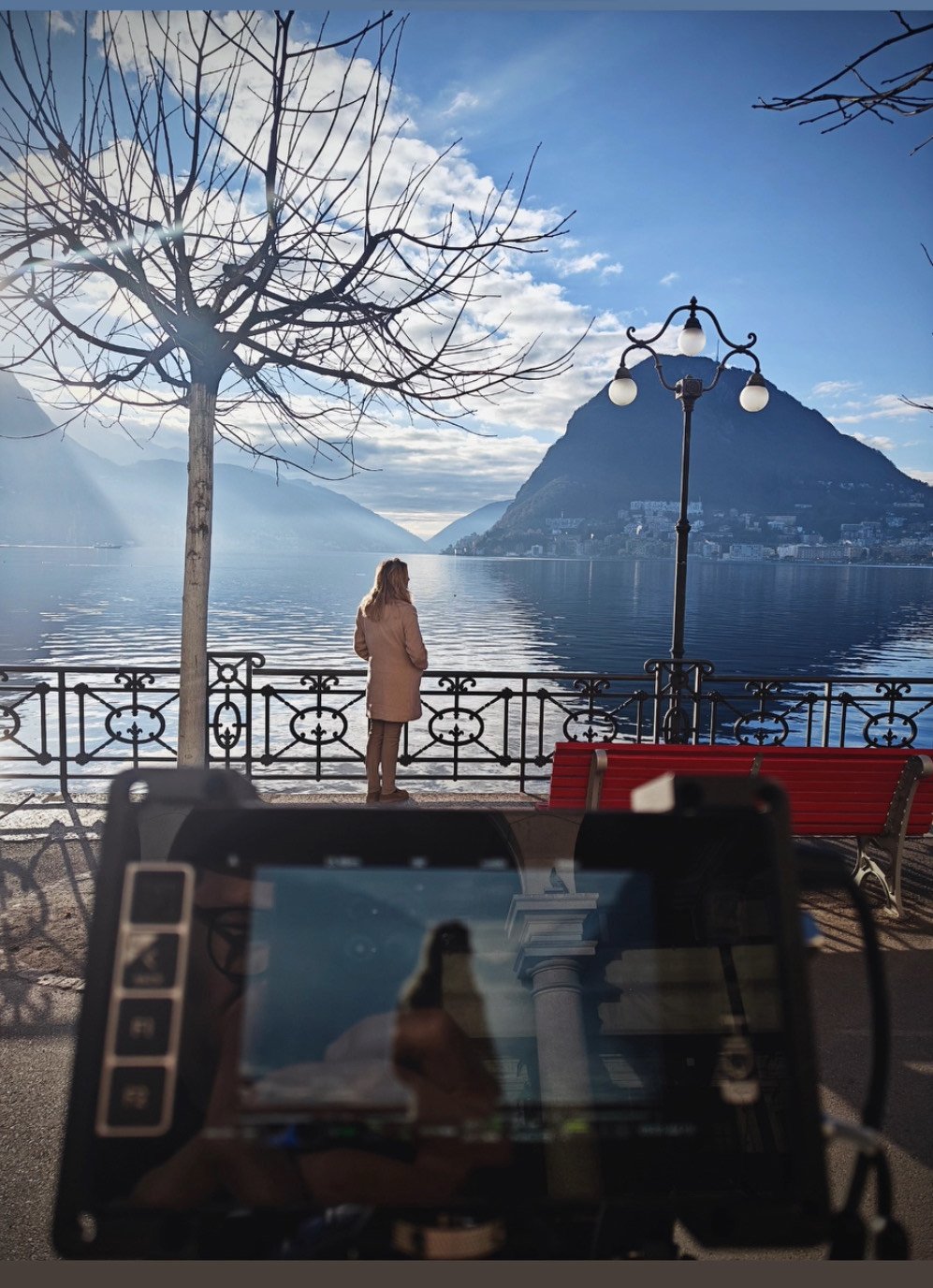 Reflections of Gauguin - Film makinh of photo. A woman dressed in a beige coat standing near a lake with mountains in the background, taking photos of the scenery. There is a bare tree, a red park bench,.