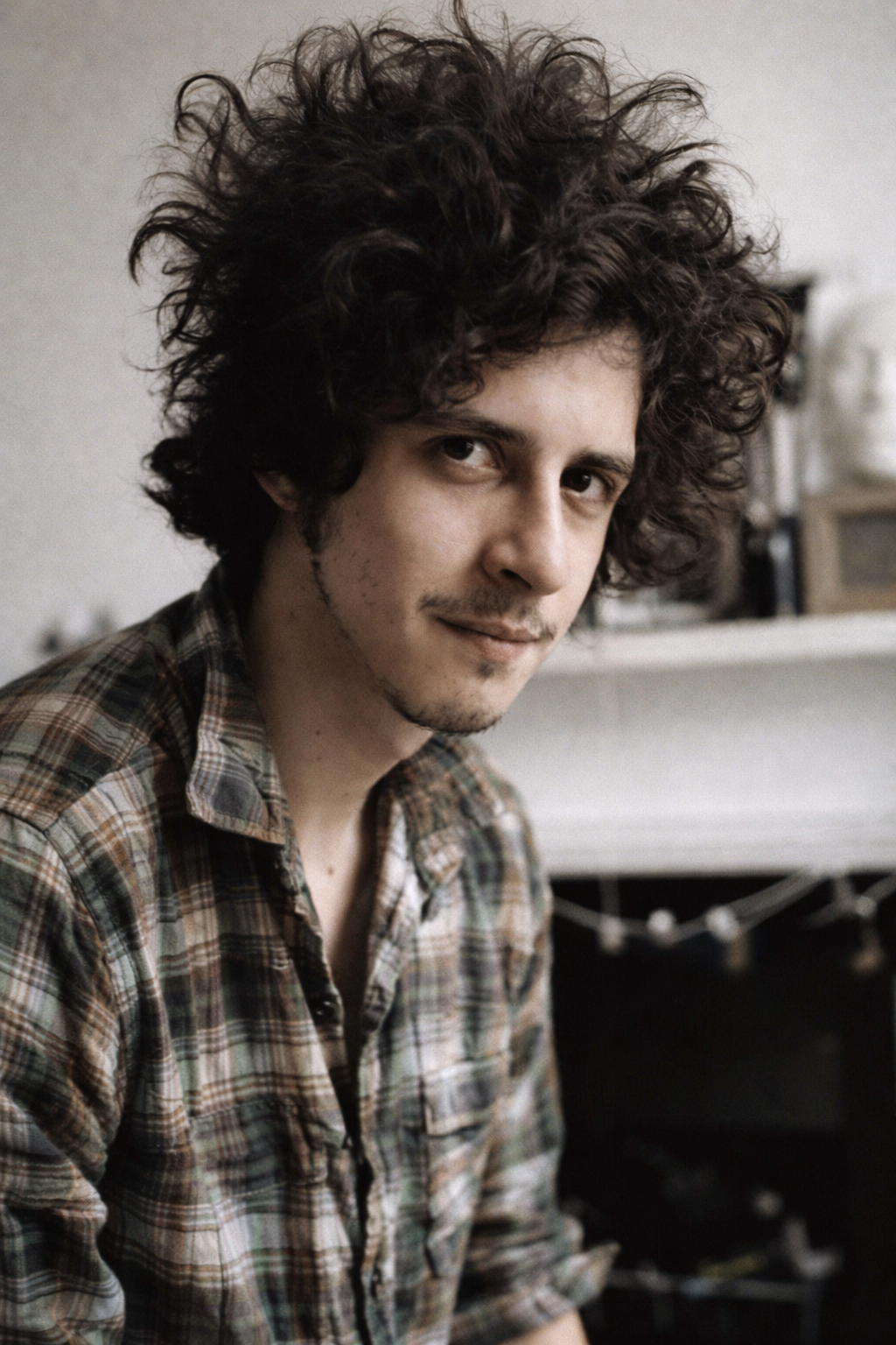 A young man with curly dark hair, a slight mustache, and light skin, wearing a plaid shirt, looking at the camera with a subtle smile in a casual setting.