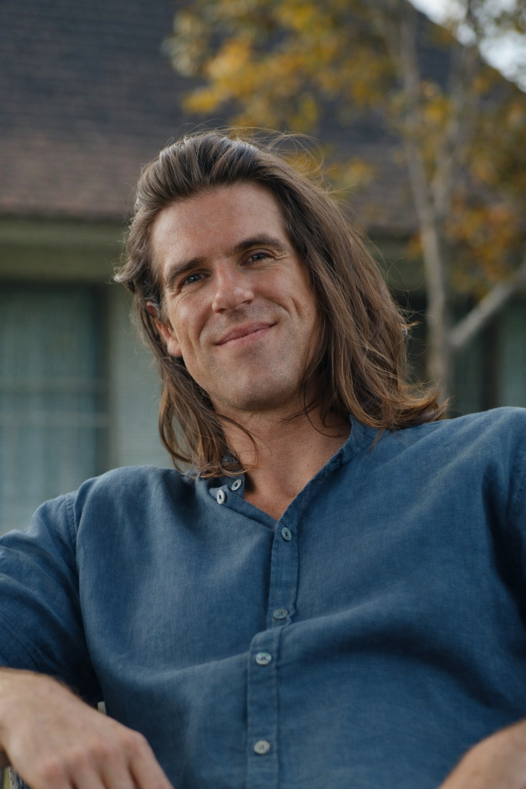 A man with shoulder-length brown hair smiling outdoors with trees and a house in the background.