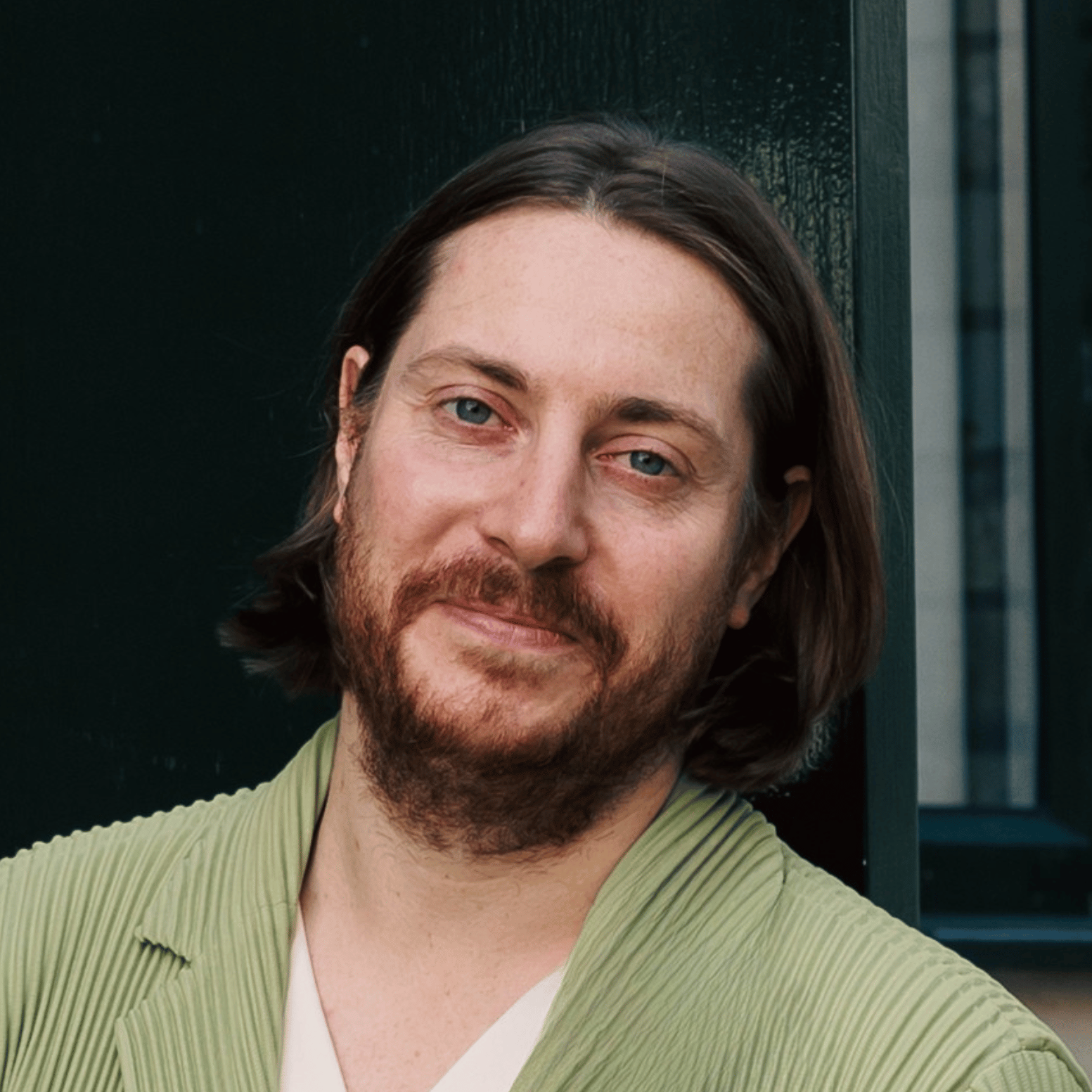 A man with shoulder-length brown hair and a beard, wearing a light green jacket, standing outdoors near a dark wall.