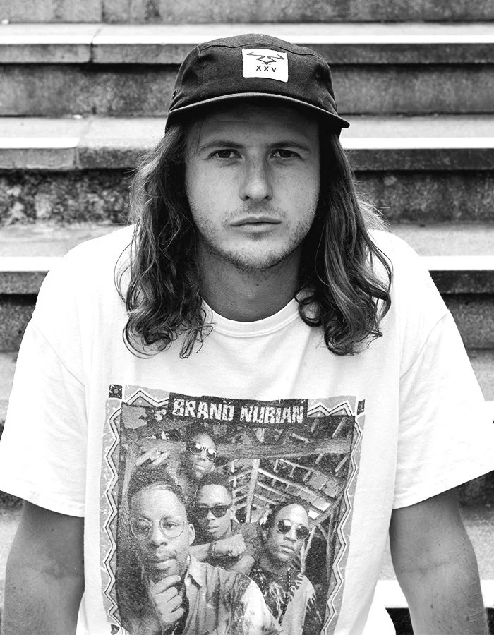 Young man with long hair wearing a black cap and a T-shirt, sitting on outdoor steps.