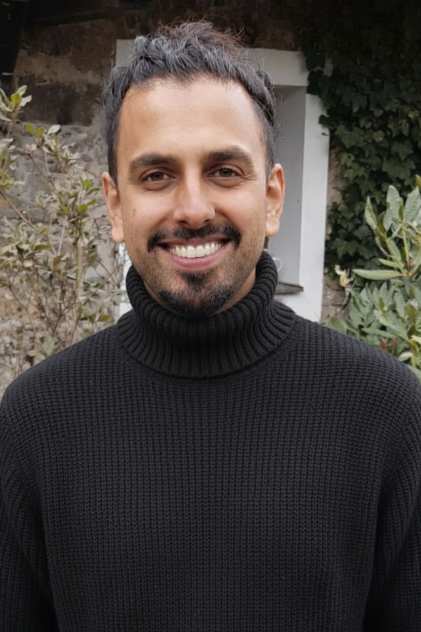 A smiling man with short dark hair and a beard, wearing a black turtleneck sweater, standing outdoors with plants and a stone wall in the background.