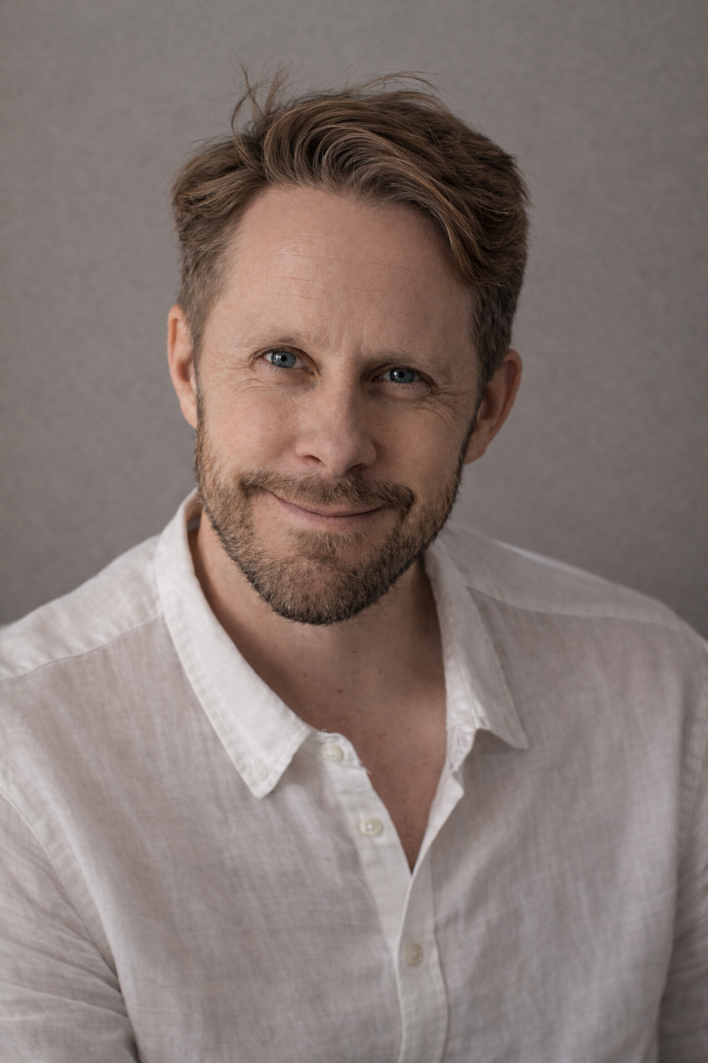 A man with light skin, brown hair and blue eyes, smiling while wearing a white button-up shirt.