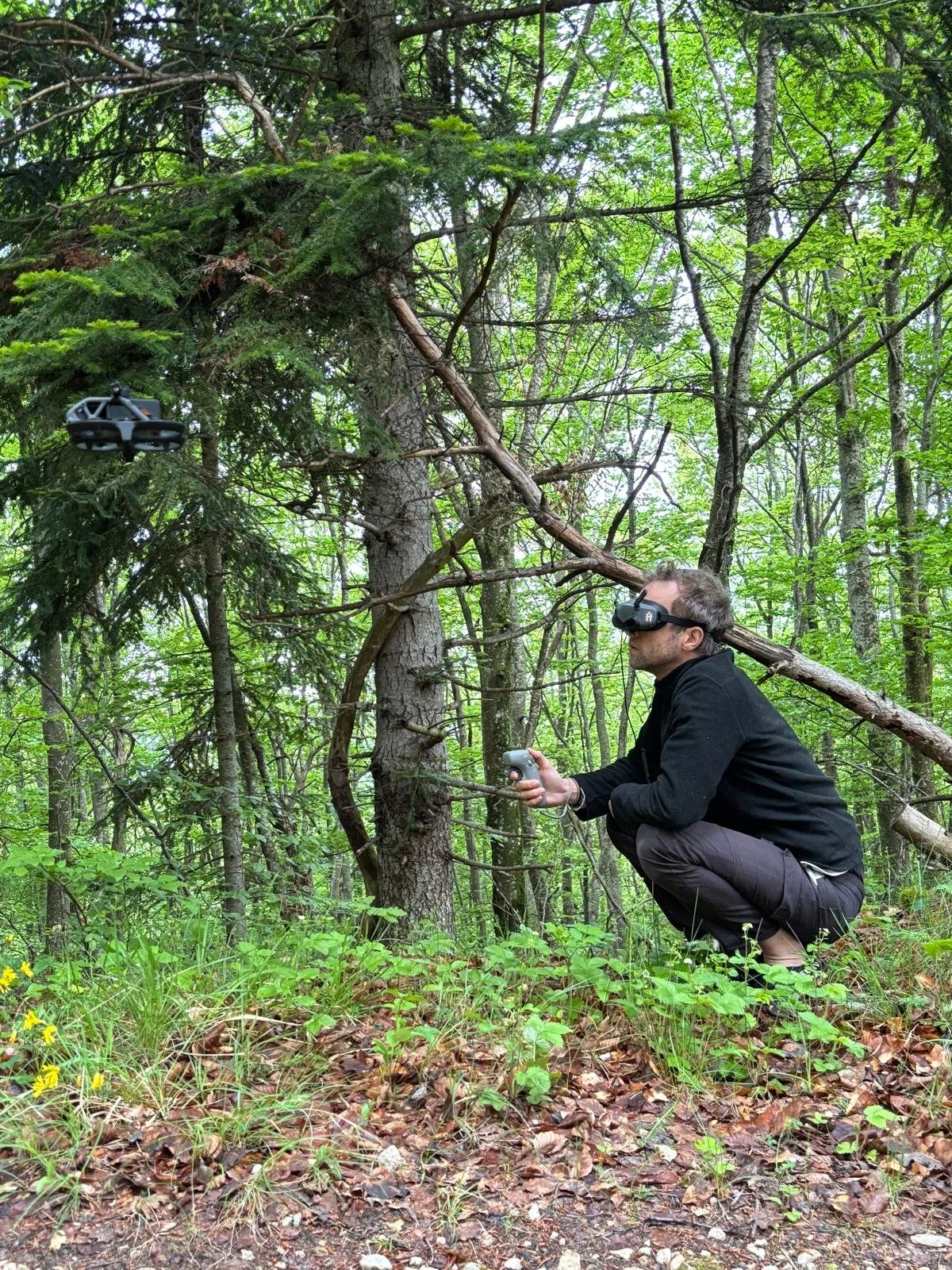 Homme utilisant un drone dans une forêt, portant des lunettes de réalité virtuelle et un contrôleur, ayant un arbre à coté de lui.