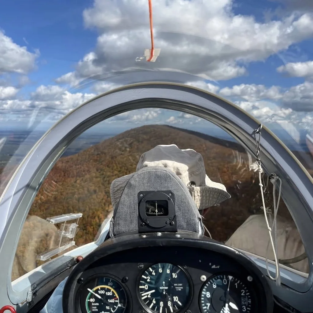View from the cockpit of a glider flying over the Catskill mountain landscape with autumn foliage, blue sky, and puffy white clouds.