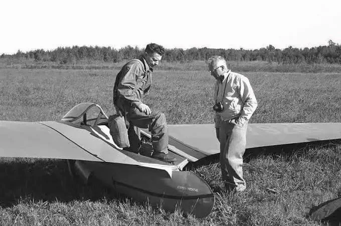 Two men around a small experimental glider on a grassy field, with one sitting on the aircraft and the other holding a camera, in black and white.