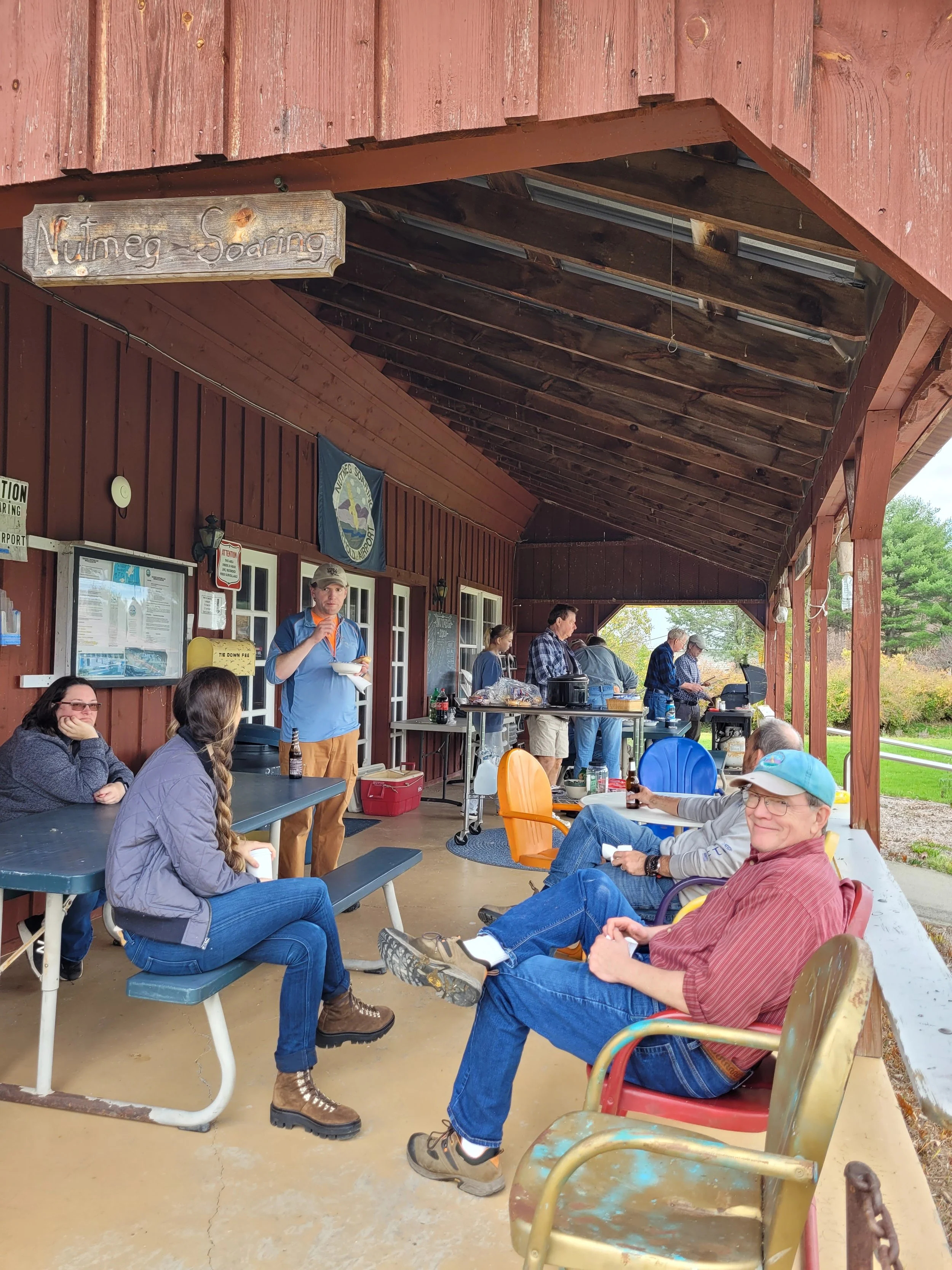 Nutmeg members gathered on the covered porch of the clubhouse, some sitting at picnic tables, others standing near a barbecue grill, enjoying outdoor food and drinks.