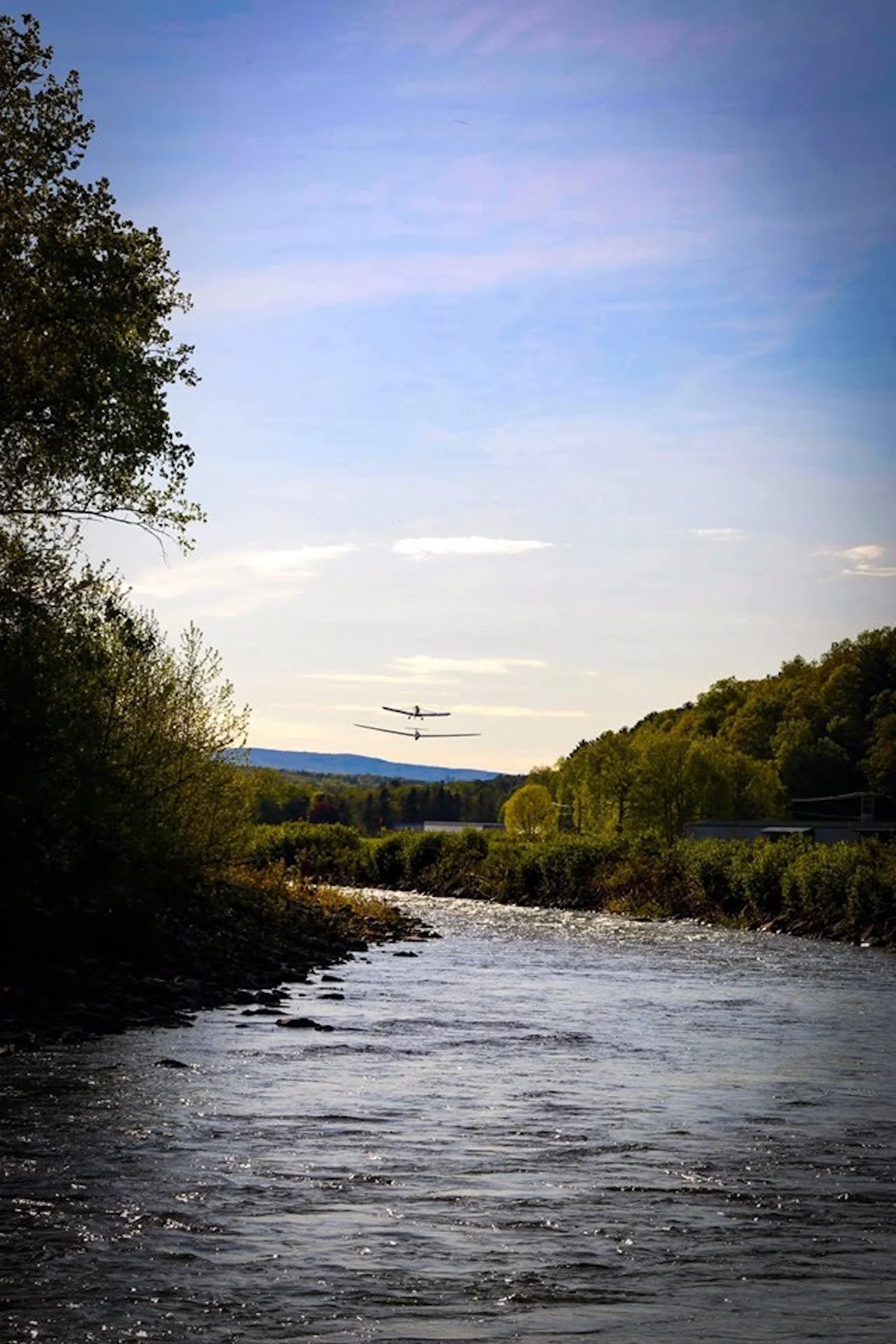 A glider being towed flying in the sky over a river with trees and hills in the background.