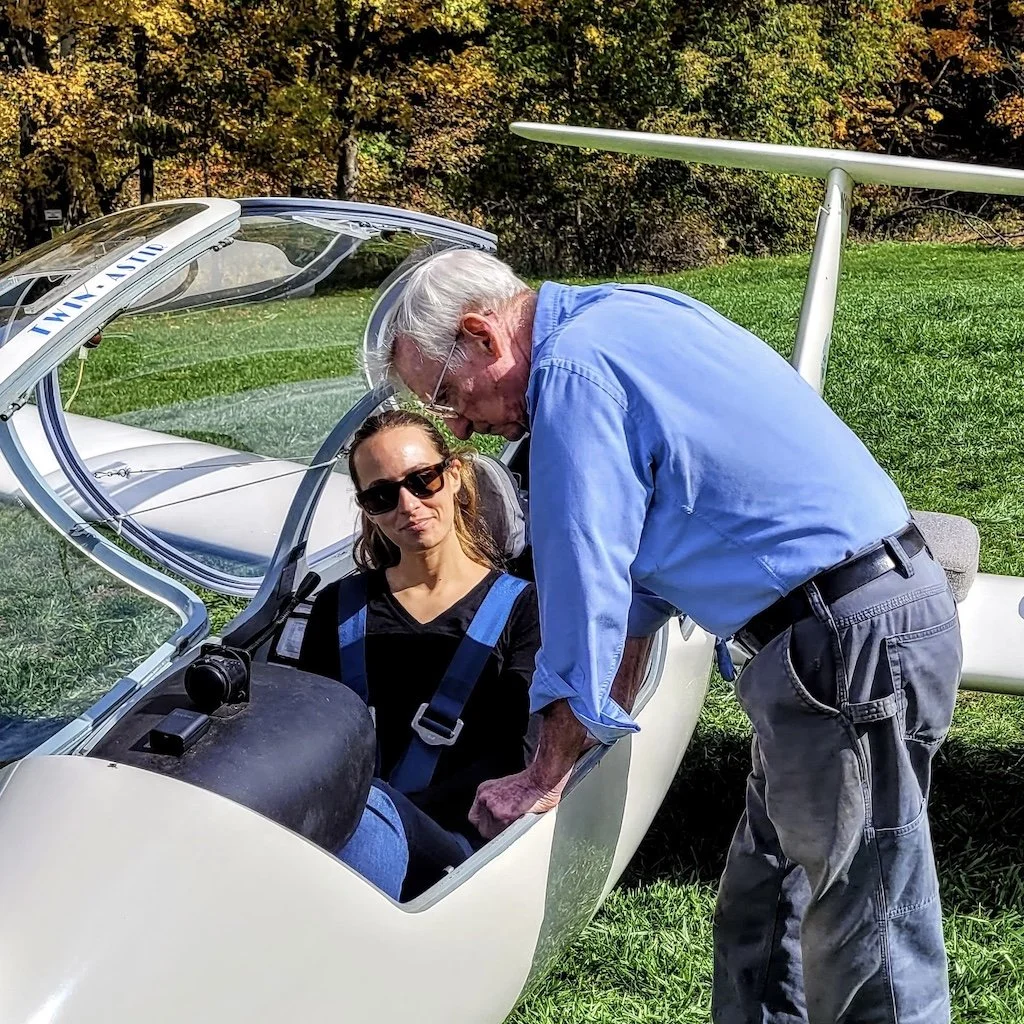 Randol is talking to a Taylor who is sitting in a white glider on a grassy field with trees in the background; the woman is wearing sunglasses and a black top.