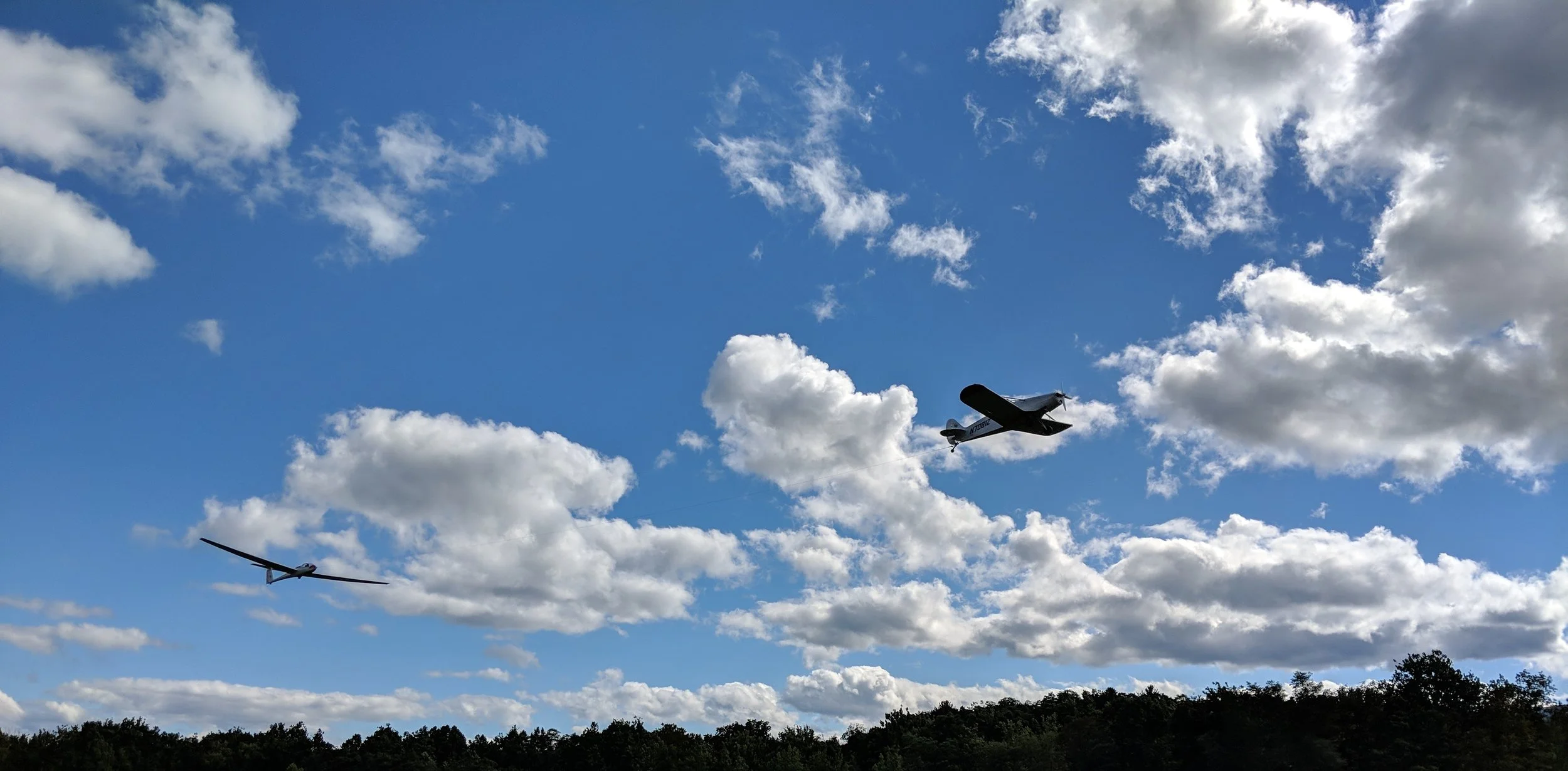 A glider being towed by an airplane flying in a partly cloudy sky above a freehold airport.