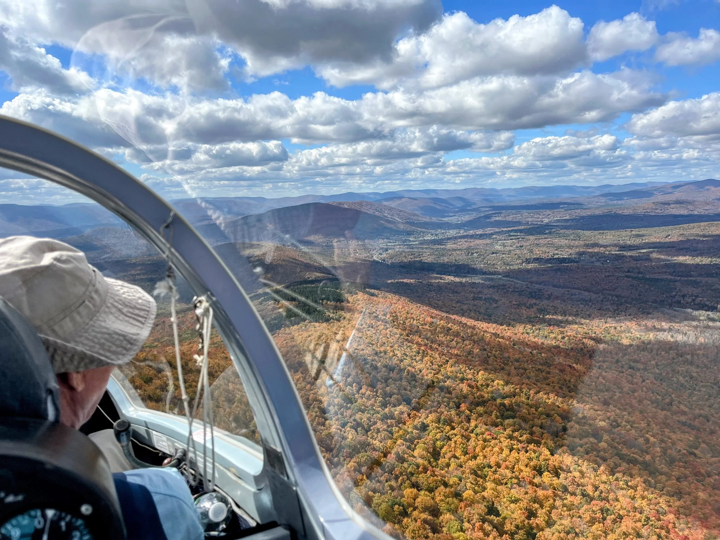 A Nutmeg member wearing a beige hat flying in a small sailplane above the Catskill mountain landscape with autumn-colored trees and a partly cloudy sky.