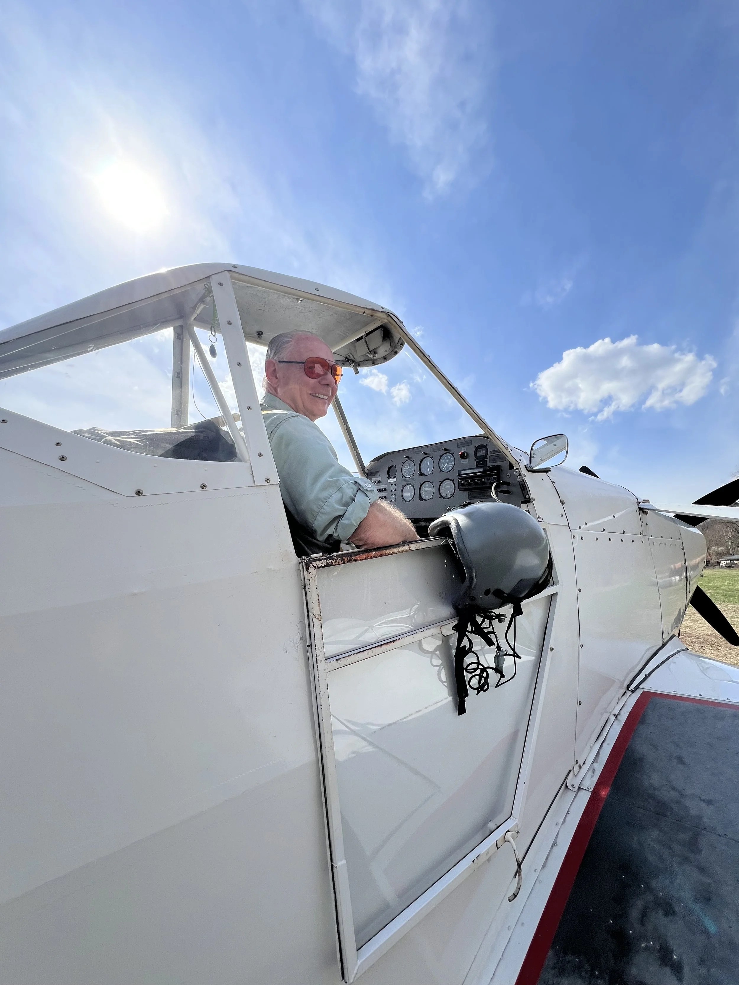 Mike with red sunglasses smiling while sitting in the cockpit of a small white airplane under a bright blue sky with some clouds.
