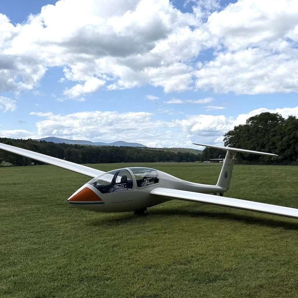 A sailplane parked on a grassy field with a cloudy sky and distant trees and mountains in the background.