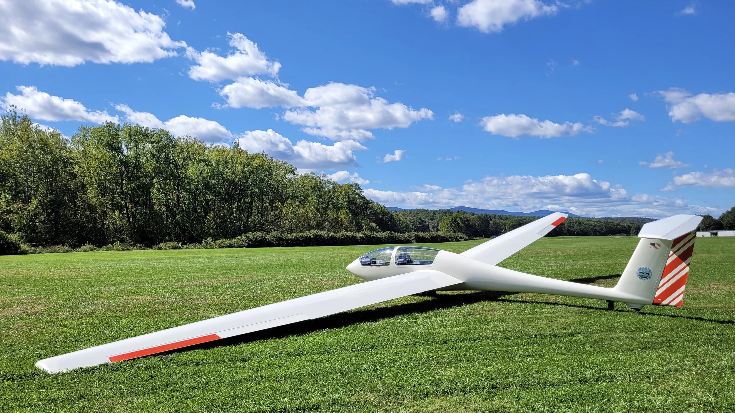 A white sailplane with red accents sitting on a grassy field under a bright blue sky with white clouds, surrounded by trees and distant mountains.