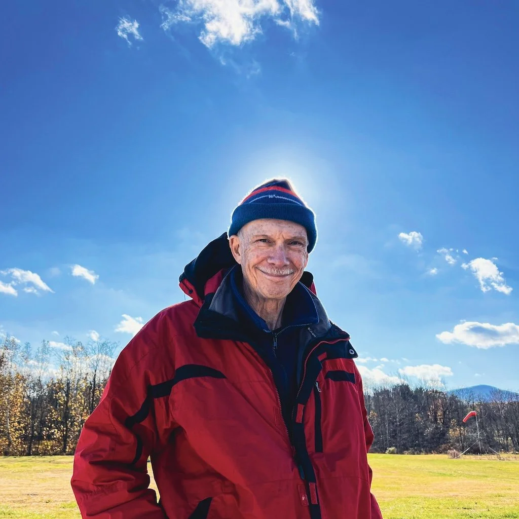 Marty wearing a red jacket and a colorful knit hat smiles outdoors on a sunny day with blue skies, scattered clouds, and a grassy field with trees and a windsock in the background.