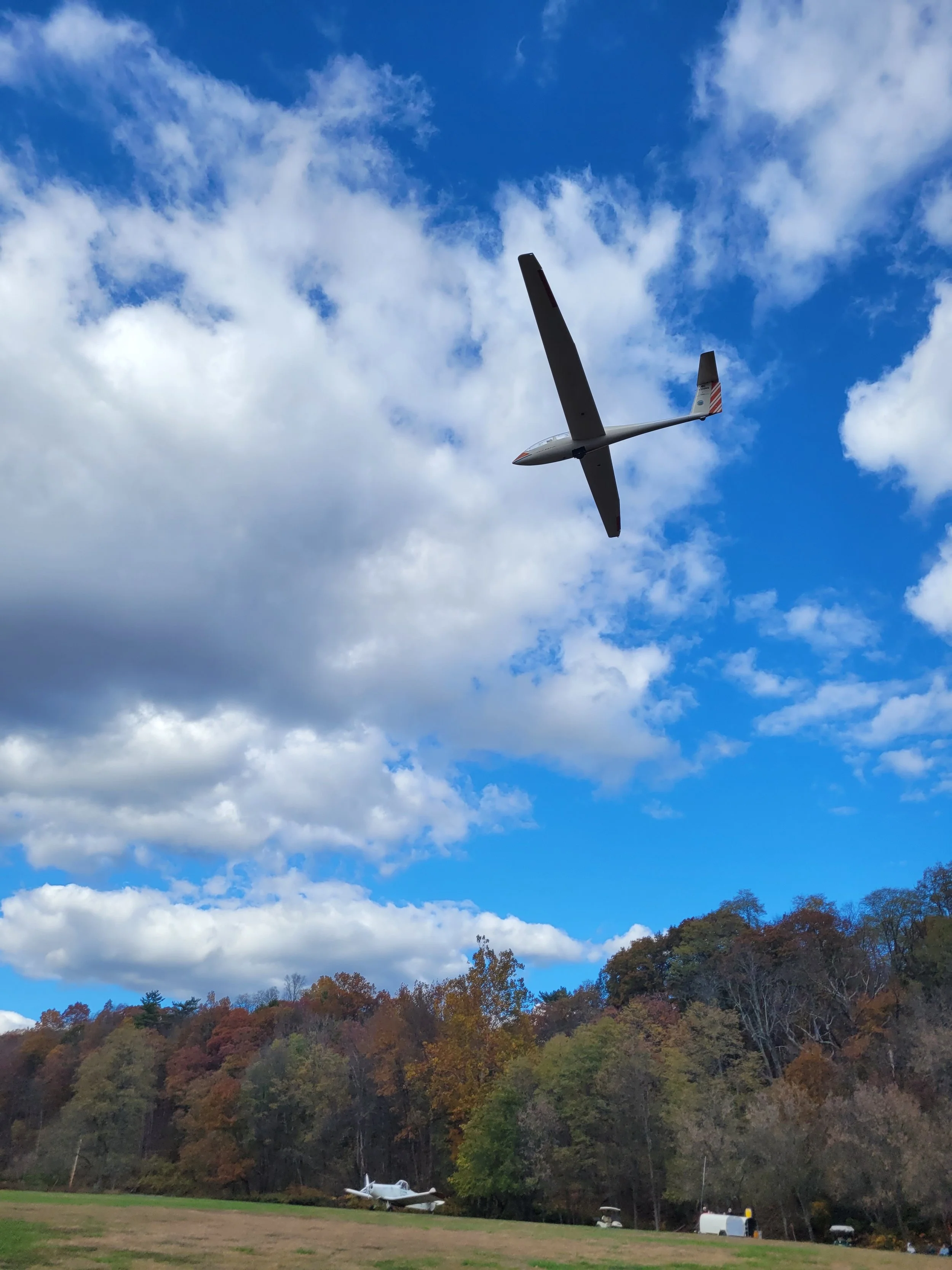 A glider on final approach flying in a partly cloudy sky with trees displaying fall colors and tow plane in the ground.