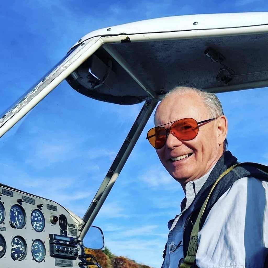 Mike is wearing sunglasses sitting in the cockpit of a tow plane, with clear blue skies in the background.