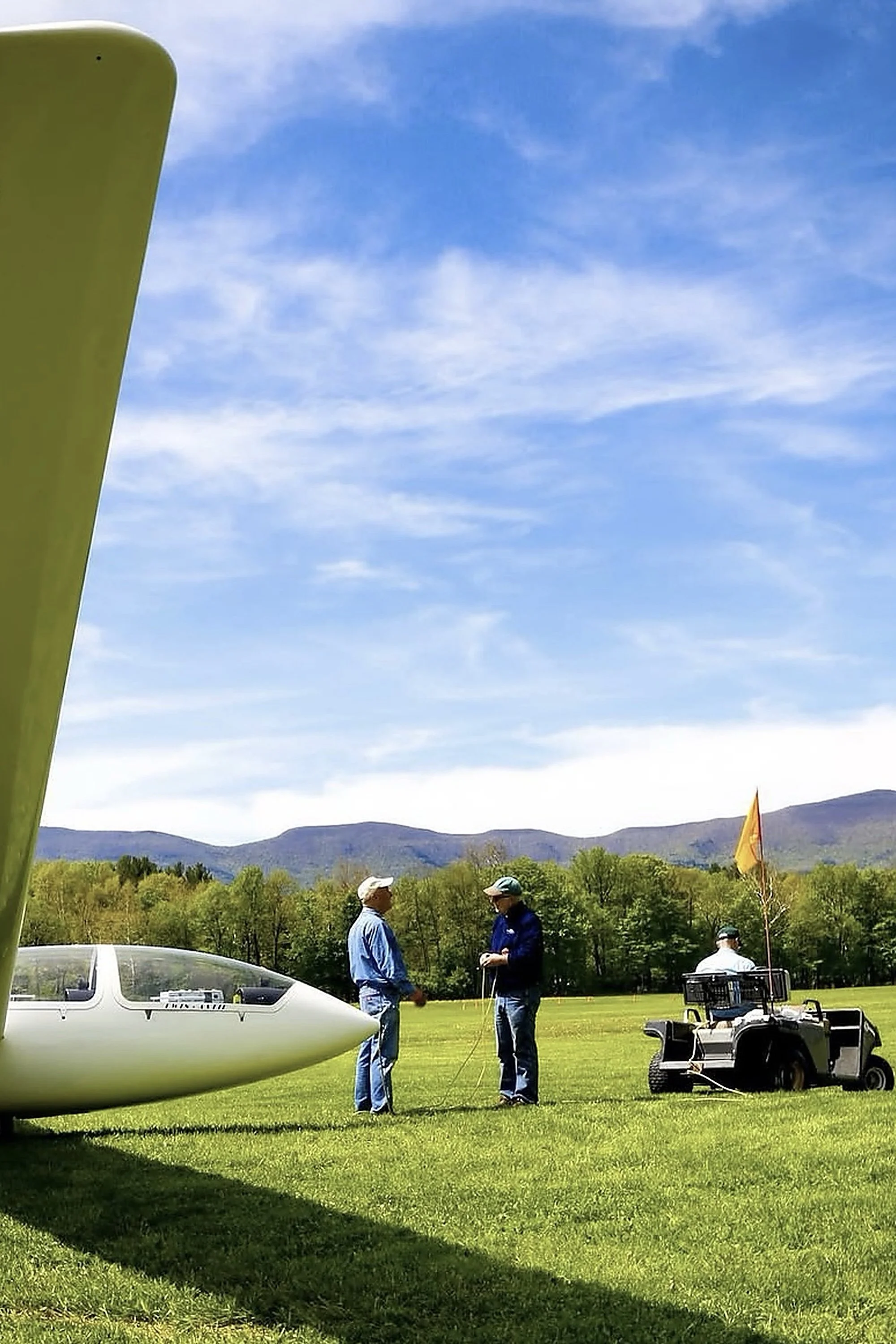 Two Nutmeg members standing on a grassy field, engaging in conversation; a member with a golf cart with a flag is nearby; part of a sailplane is visible on the left side; mountains and trees are in the background under a partly cloudy sky.
